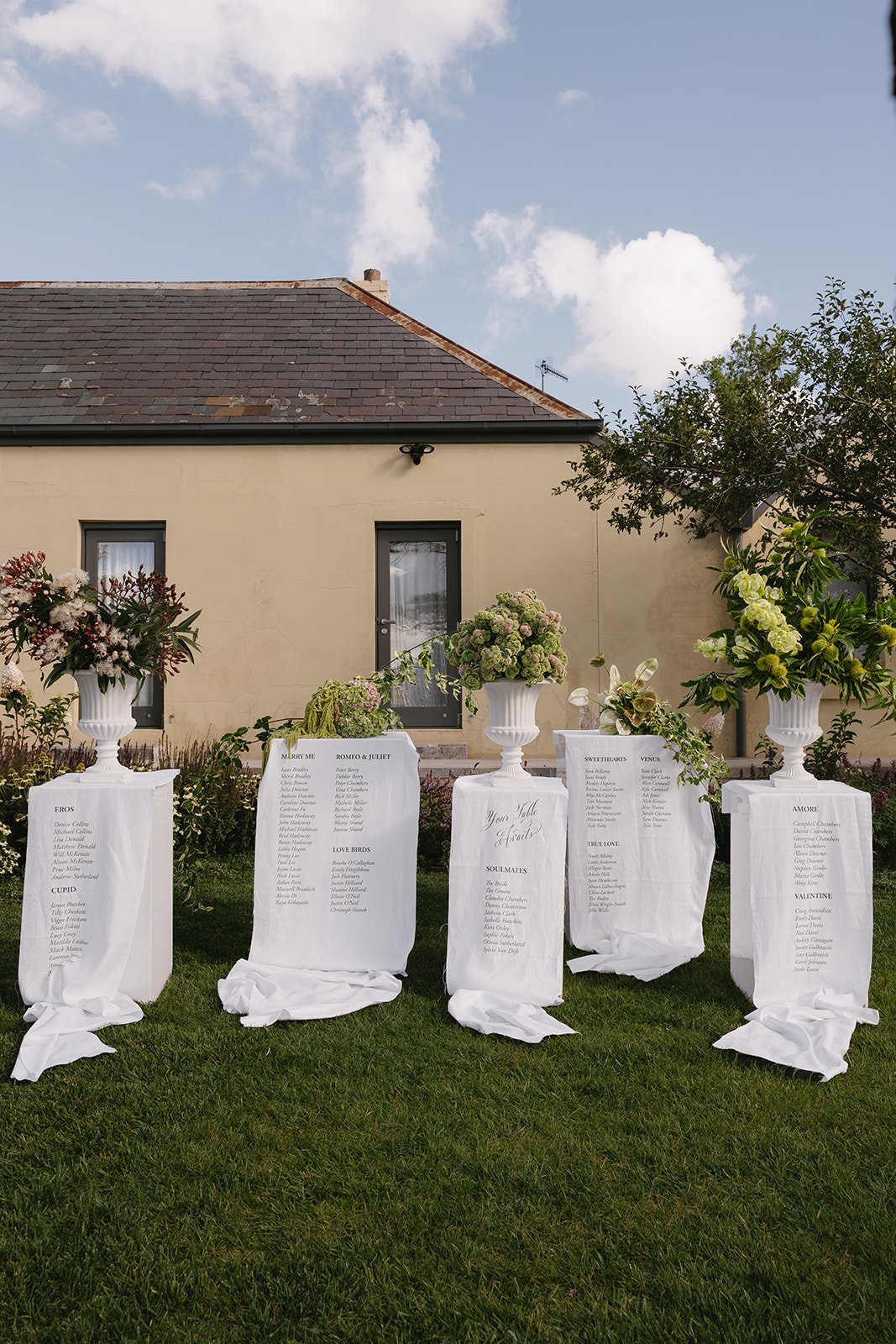Outdoor wedding seating chart display with large white vases of flowers on pedestals, draped with white cloth, set against a beige house and green lawn, under a partly cloudy sky.