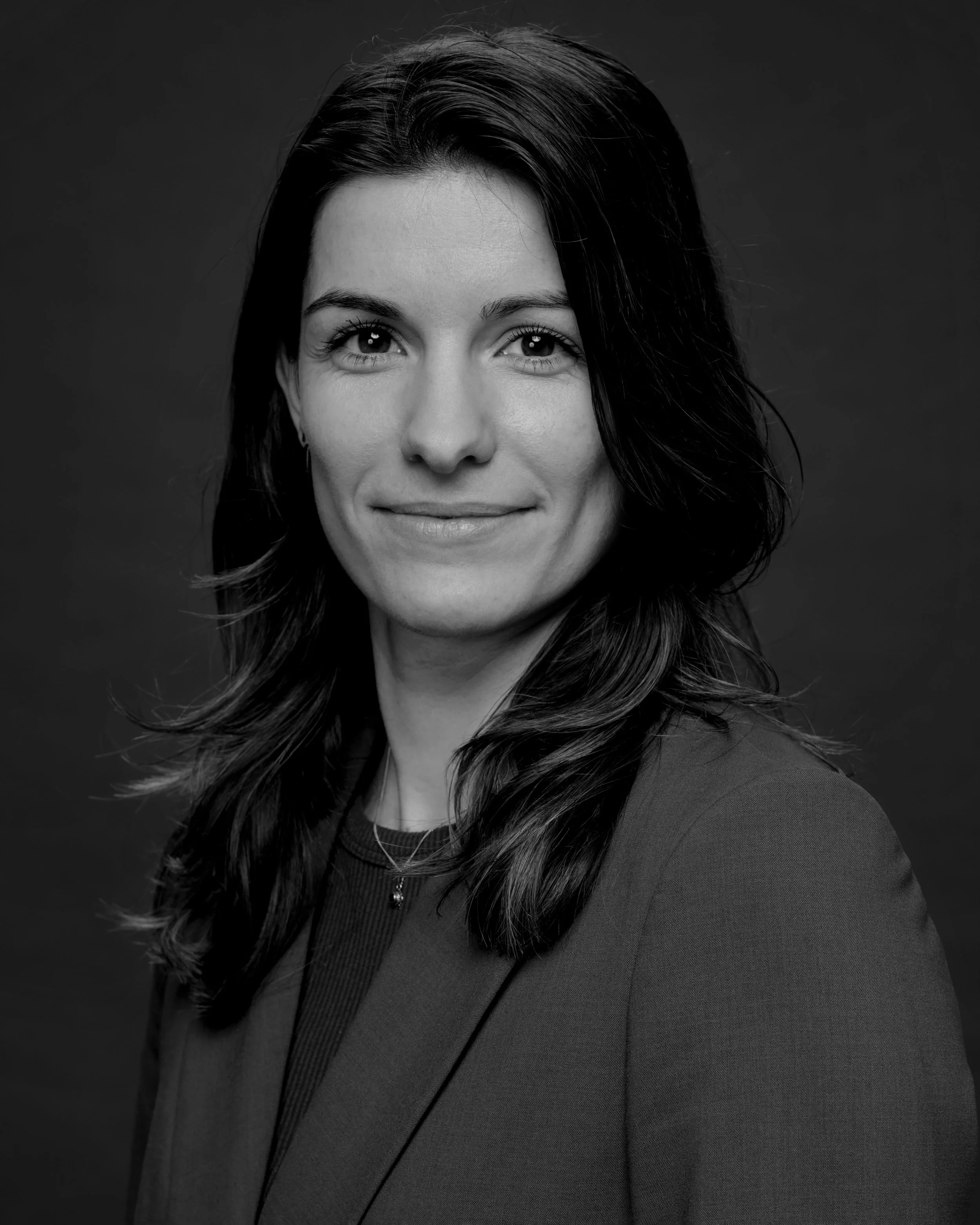 Black-and-white portrait of a smiling woman with long wavy hair, wearing a blazer and a necklace, against a dark background.