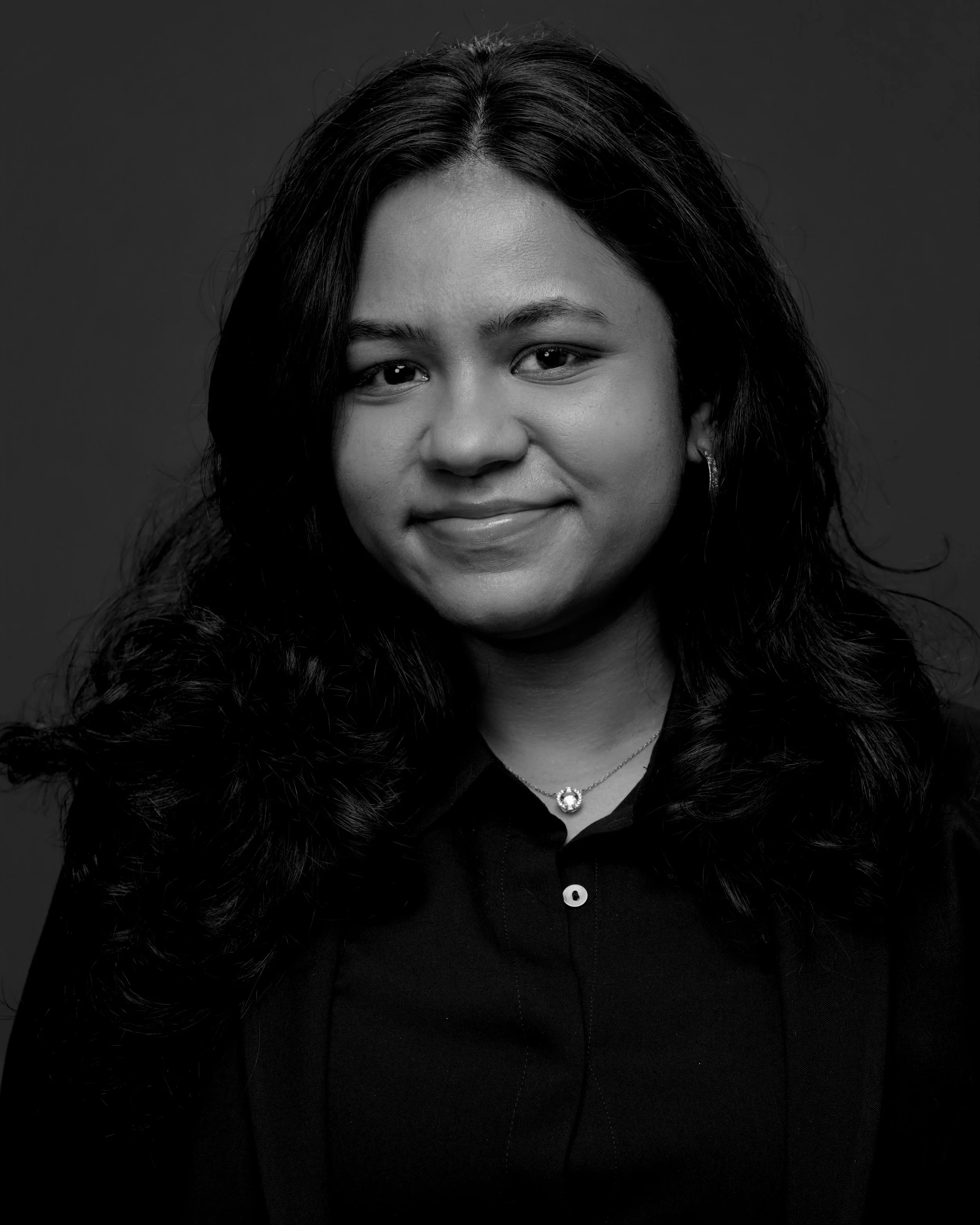 Close-up black and white photograph of a woman with medium-length dark hair, wearing a black shirt, and a necklace with a round pendant.