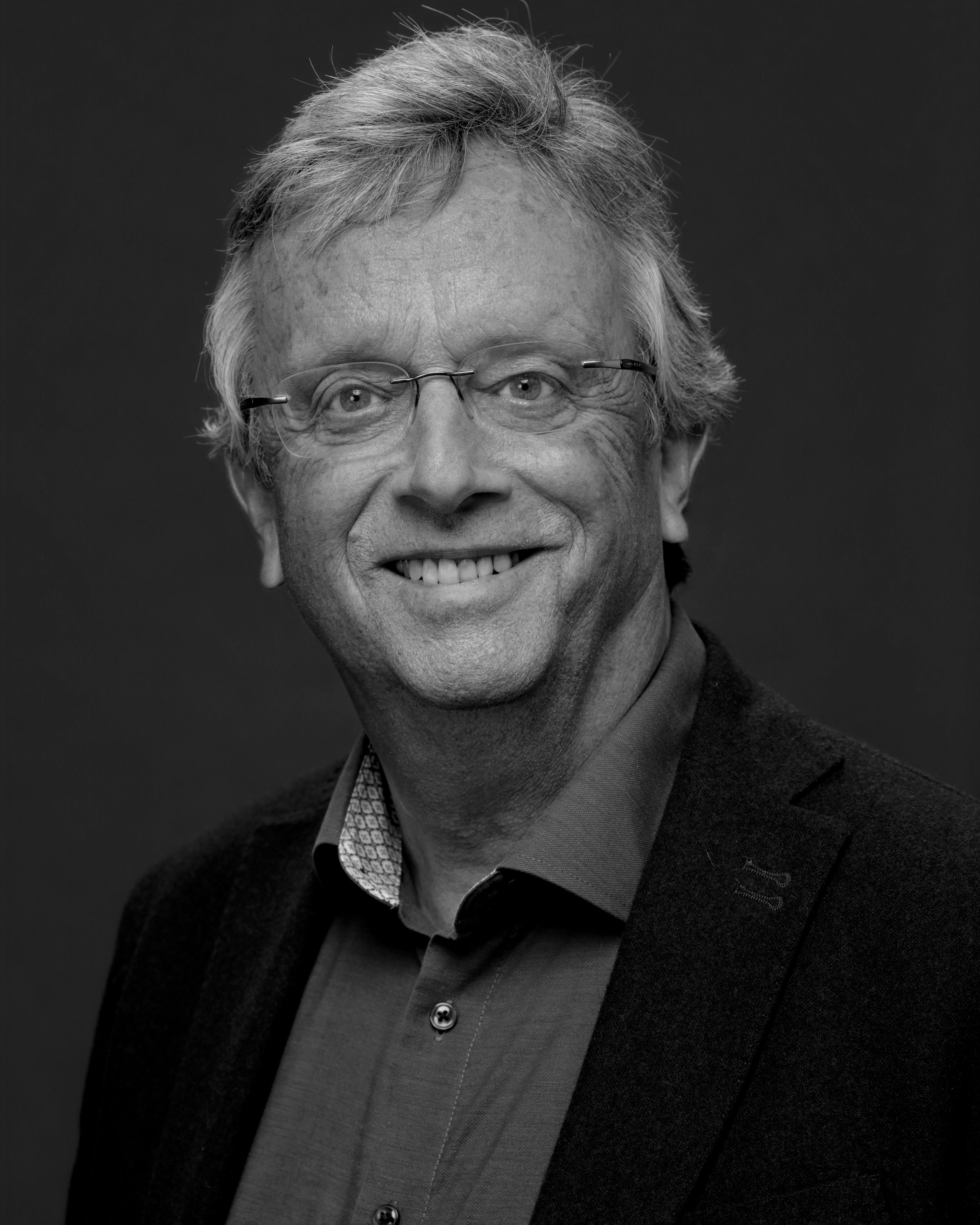 Black and white portrait of an older man with glasses, smiling, with gray hair, wearing a collared shirt and a blazer.