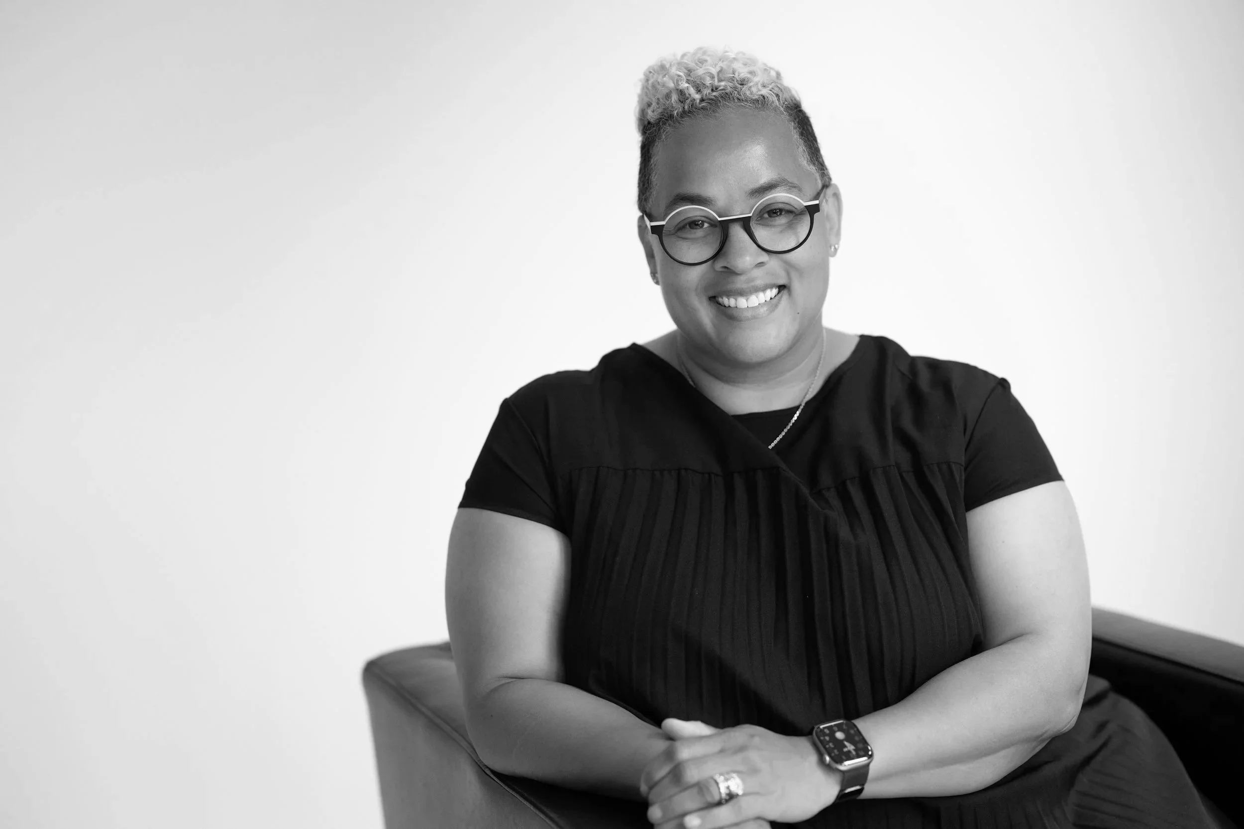 Black and white photograph of a smiling woman with glasses, short curly hair, wearing a dark dress, sitting on a chair with arms crossed, looking at the camera.