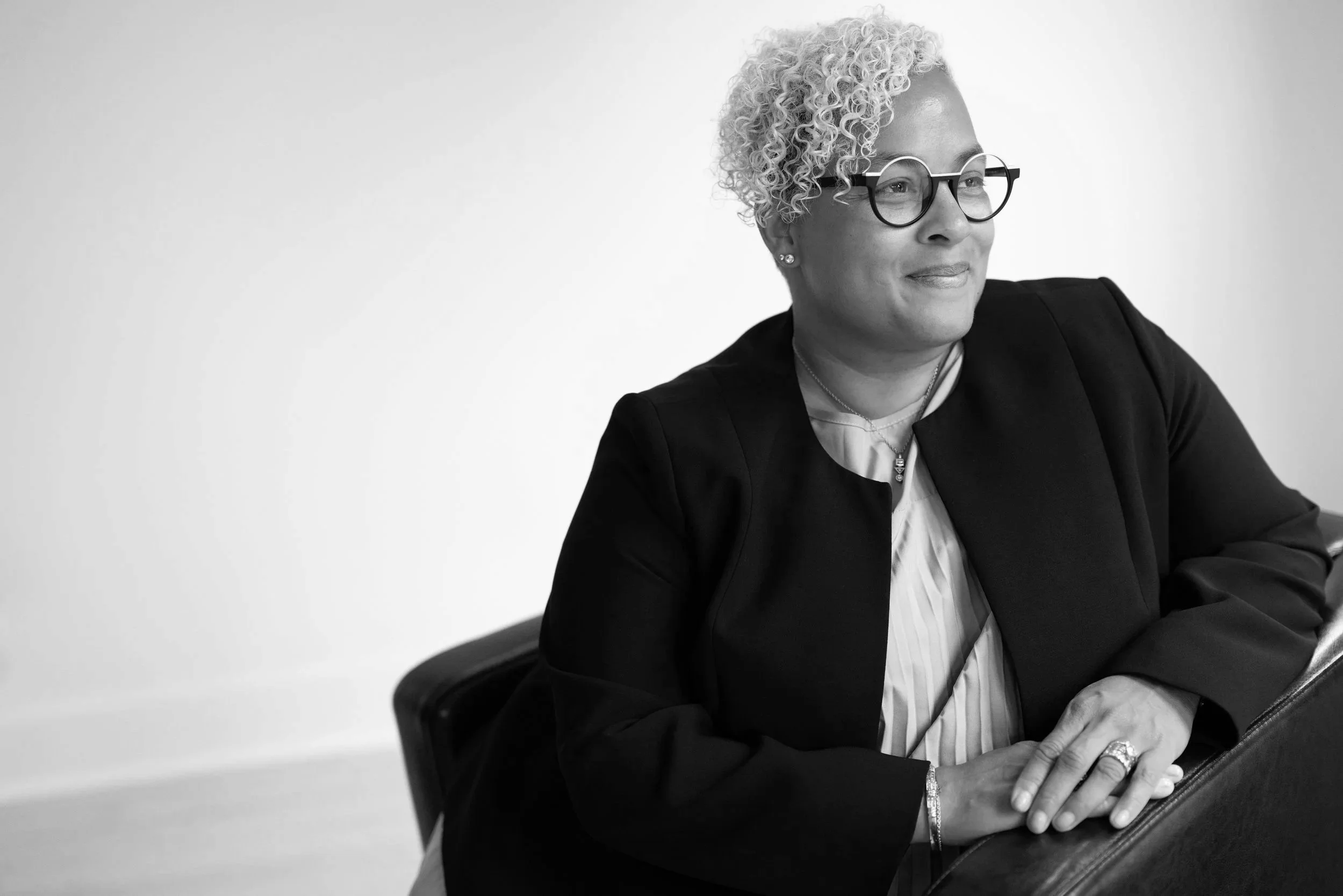 Black and white photo of a woman with short, curly gray hair wearing glasses, a light-colored blouse, and a dark blazer sitting at a desk or table with her hands clasped, looking to the right with a slight smile.