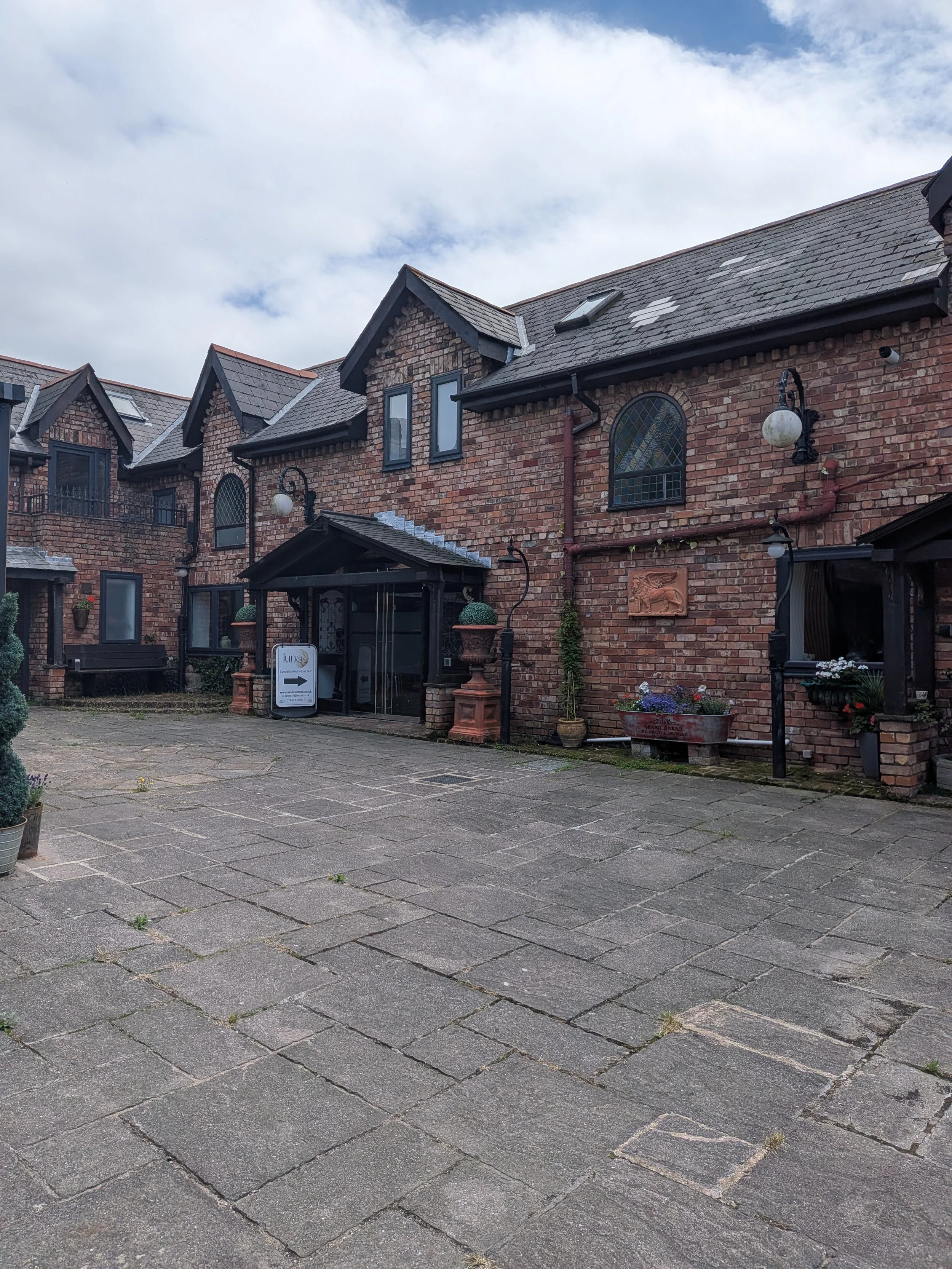A brick building with a gabled roof, decorative windows, outdoor lamps, and potted plants. The cobblestone courtyard is in the foreground under cloudy sky.