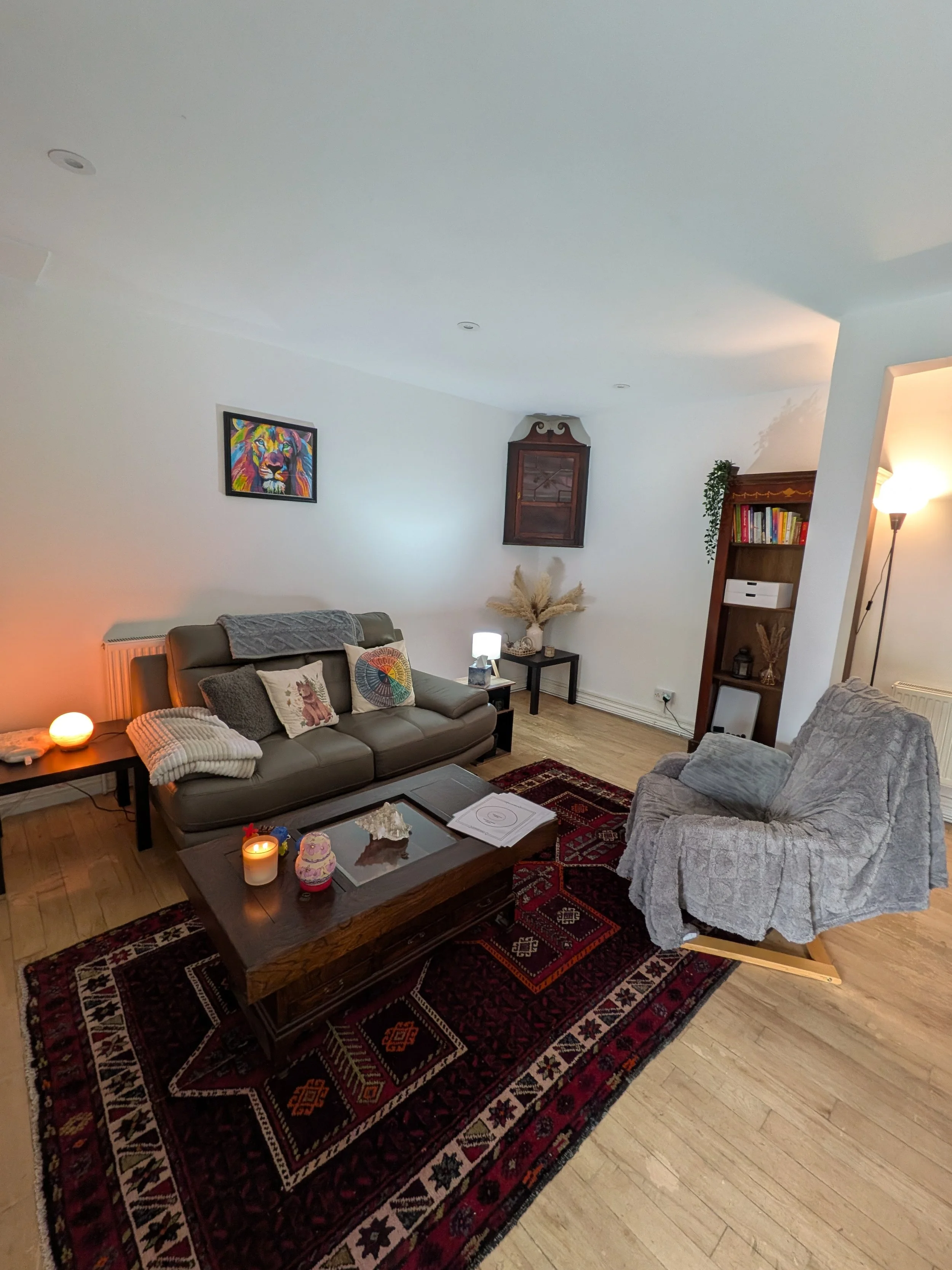 A cozy living room with a leather sofa, armchair, coffee table with candles and decorations, patterned rug, wall art, and bookshelf, illuminated by warm lighting.