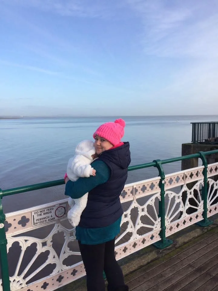 A woman in a pink knit hat and navy vest holding a small baby in a white coat on a pier by the water during daytime.
