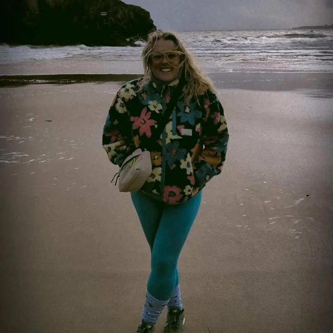 Woman standing on the beach wearing colorful fleece jacket, teal leggings, and hiking boots with a small beige bag over her shoulder, with ocean waves and cliffs in the background.