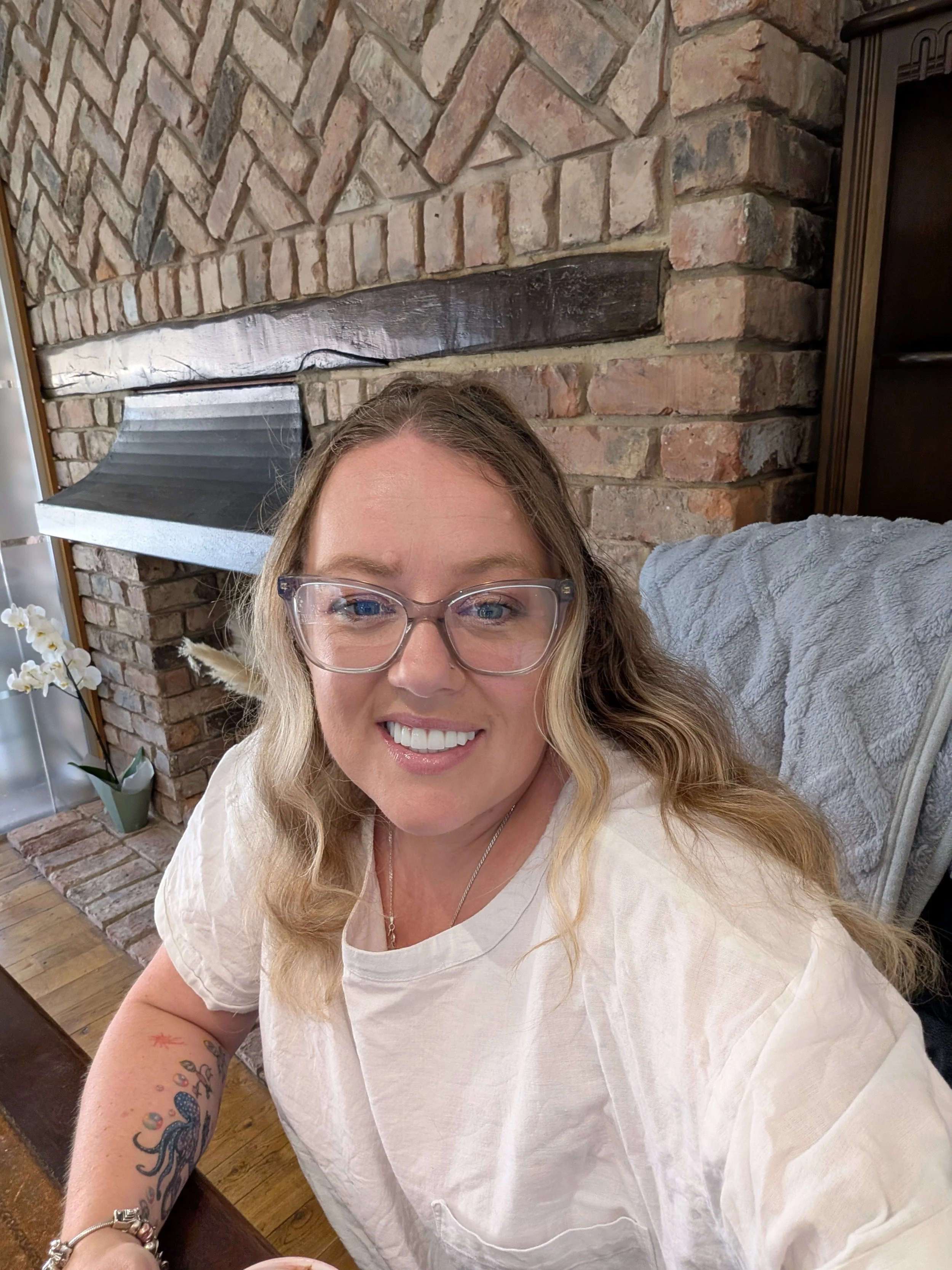 Close-up of a smiling woman with blonde hair and glasses, seated indoors in front of a brick fireplace and a wooden shelf, wearing a light-colored top, with a tattoo on her left arm.