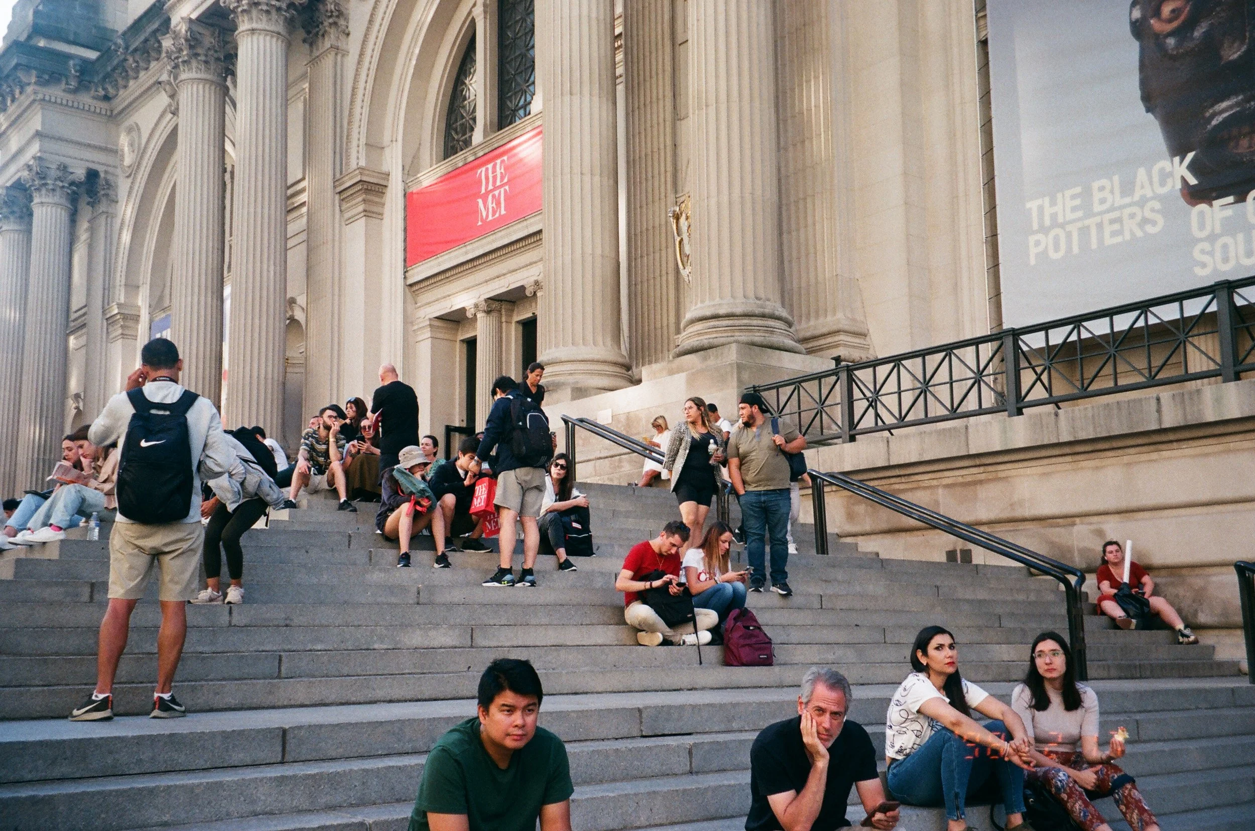 People sitting and gathering on the stairs outside a historic building with classical columns and architectural details. Some individuals are engaged in conversations, using phones, or resting.
