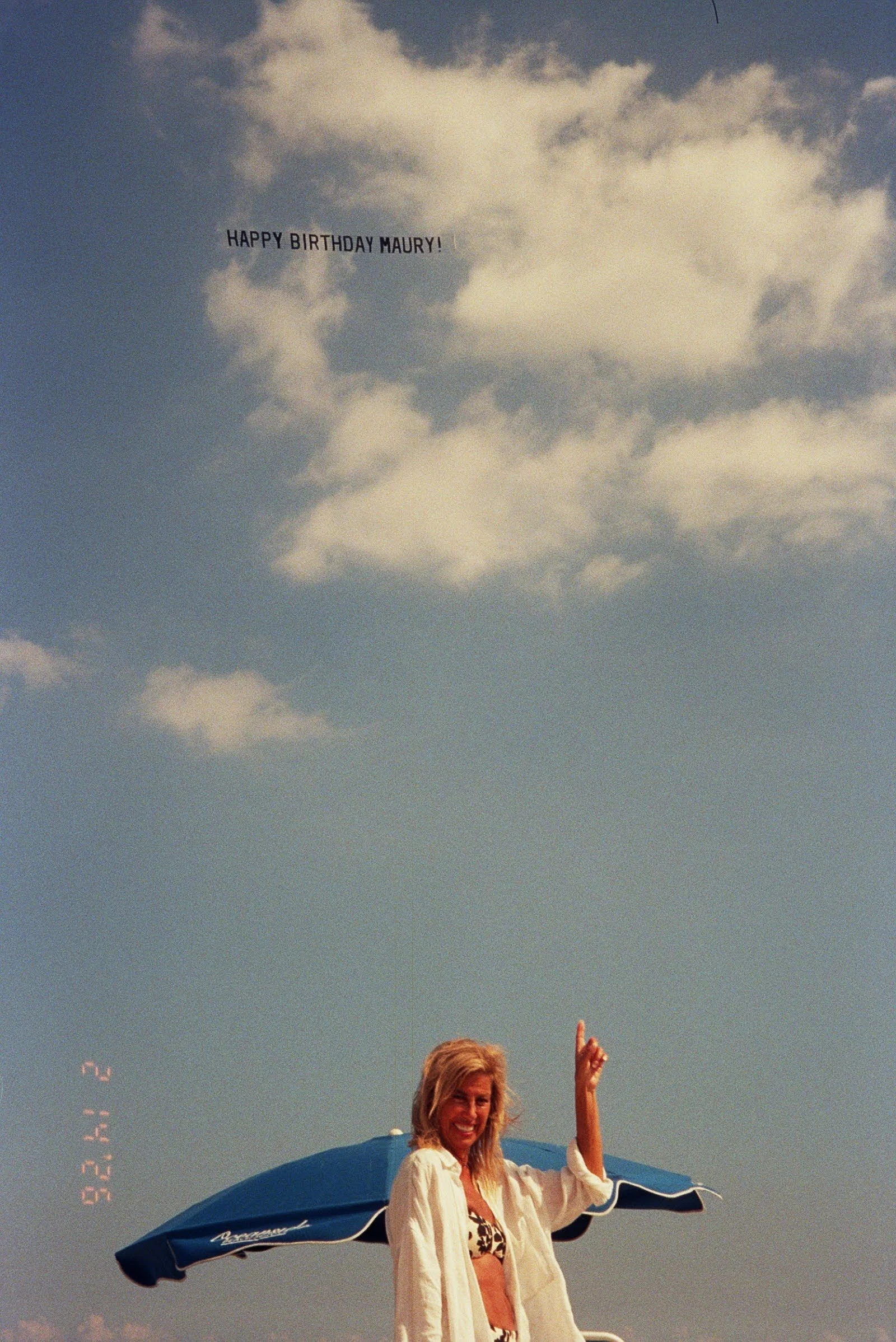 A woman with blonde hair smiling and waving under a blue beach umbrella, with a clear sky and clouds in the background.