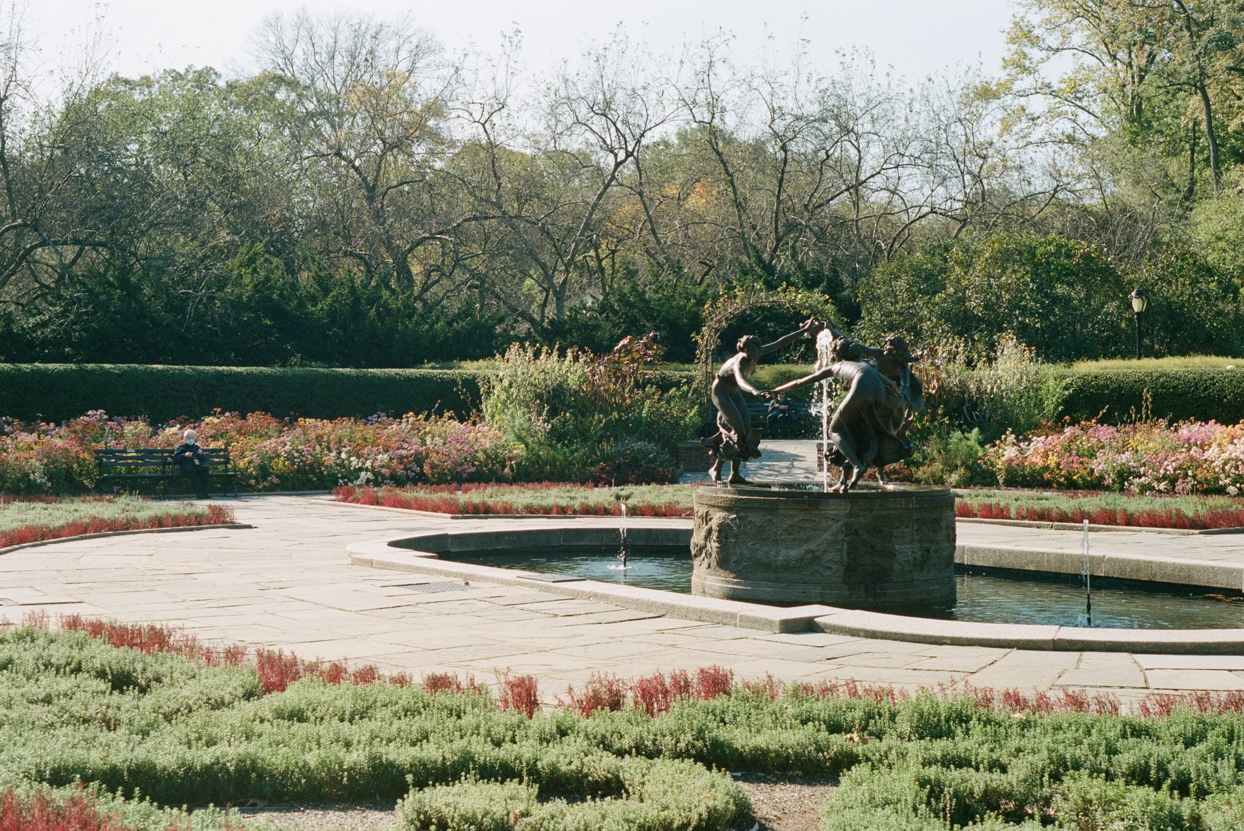 A park with a fountain featuring statues of children, a person sitting on a bench, and trees with autumn foliage.