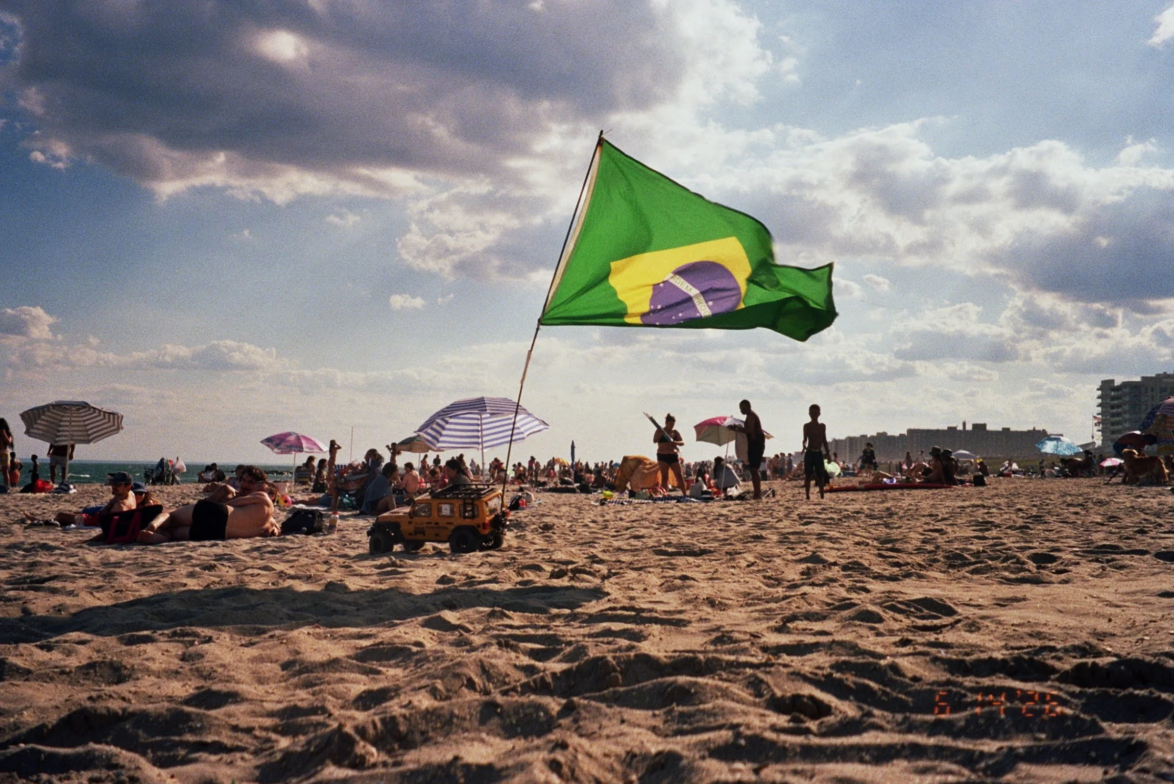 Beach scene with numerous people, umbrellas, and a large Brazilian flag on a flagpole. The sky is partly cloudy with a mix of sunlight and shadows, and some buildings are visible in the distance.