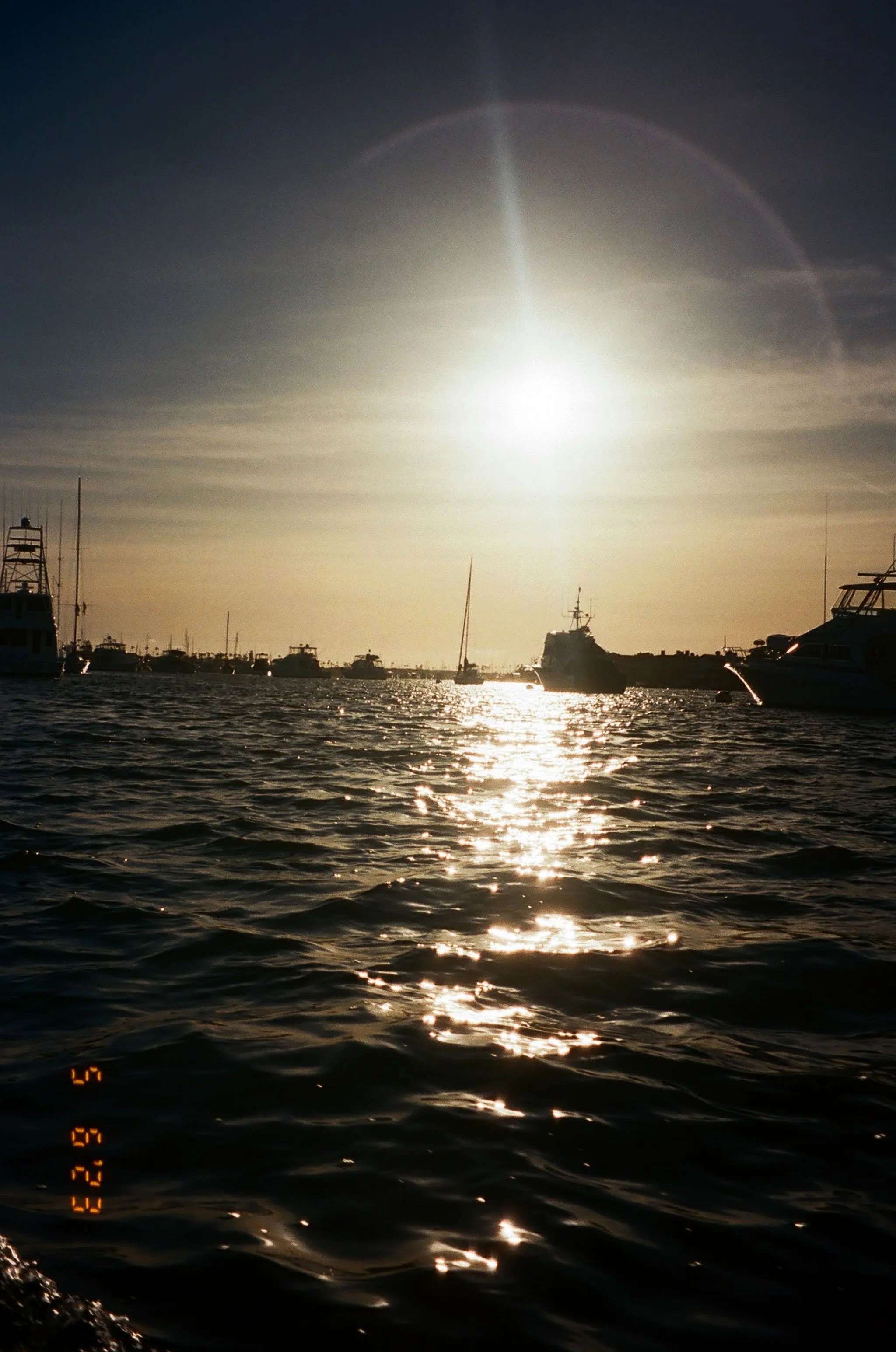 Sunset over a marina with boats and yachts on calm water, reflections on the surface, and a bright sun in the sky.