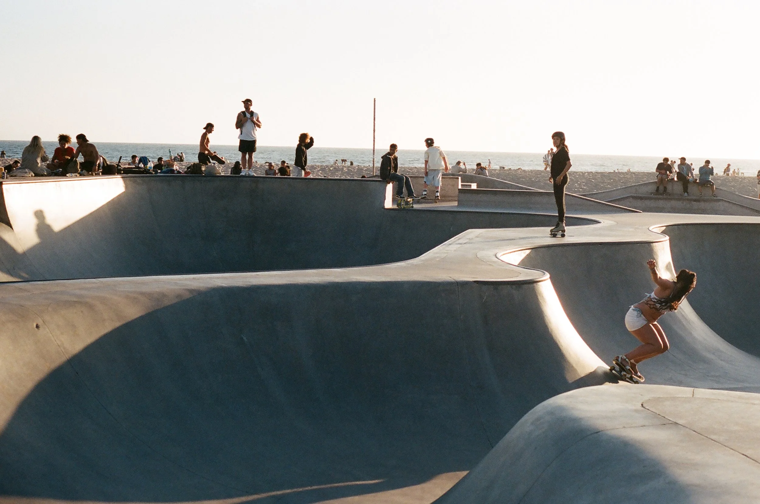 Skateboarders skate at a concrete skatepark near the beach during sunset. Many people are sitting on the sand and the skatepark.