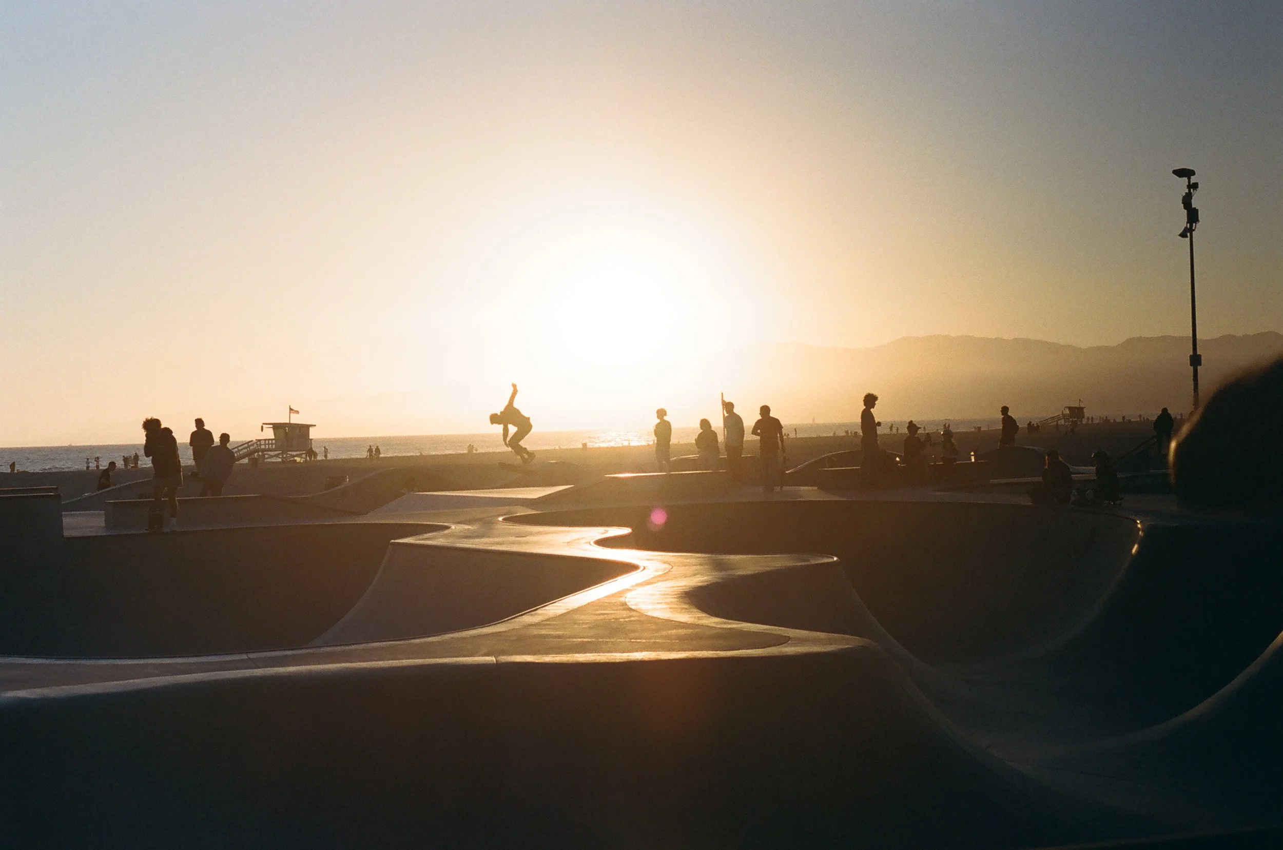 Silhouette of people at a skatepark during sunset with a beach and ocean in the background.
