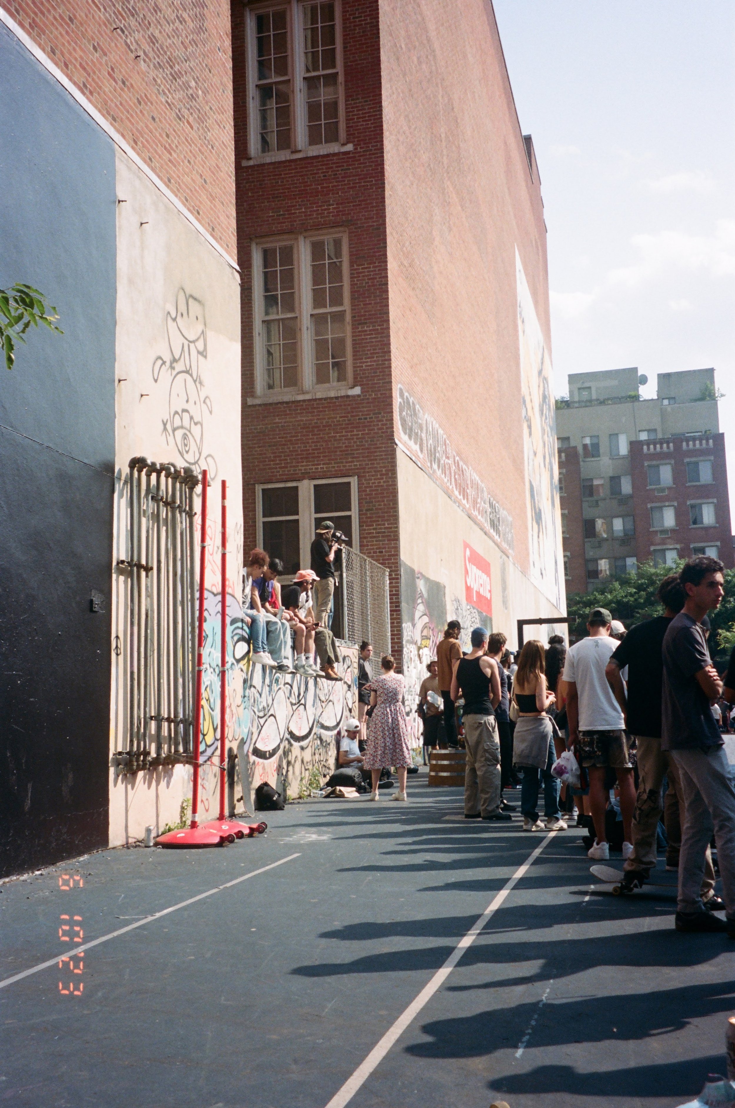 People gathered outside along a painted basketball court with a graffiti-covered wall in the background. Some individuals are sitting on the ledge, others are standing or walking. The scene is in an urban environment with tall buildings in the distan