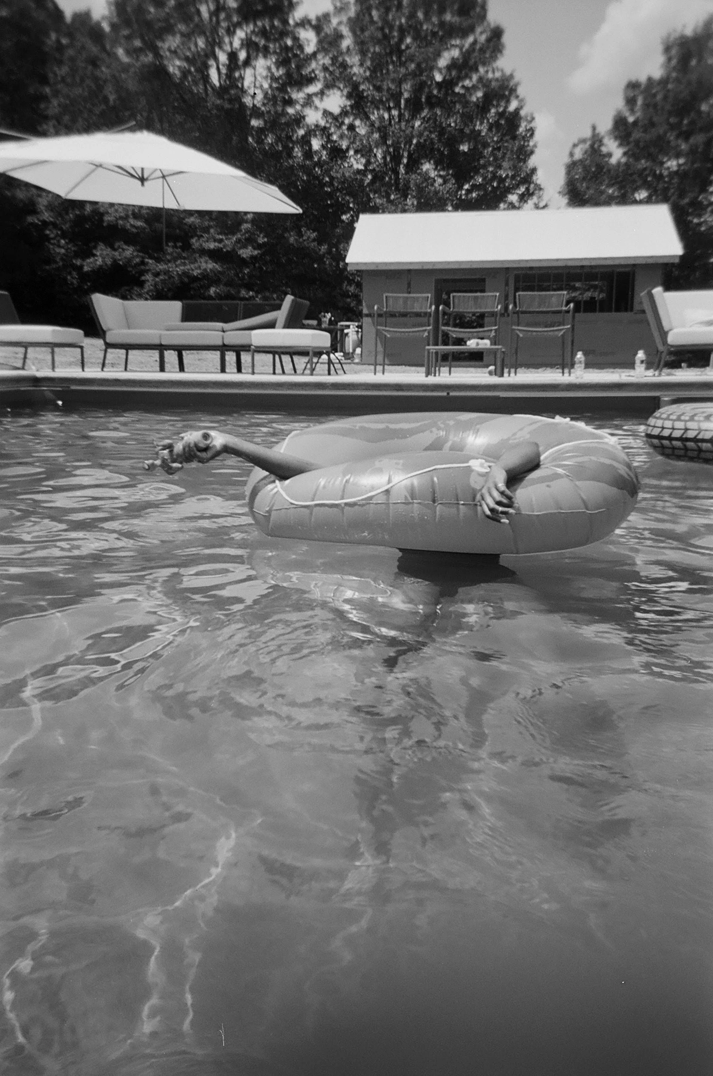 A person is floating in a swimming pool with a inflatable ring, holding a drink. Behind the pool, there are outdoor furniture and an umbrella, with trees and a small building in the background.