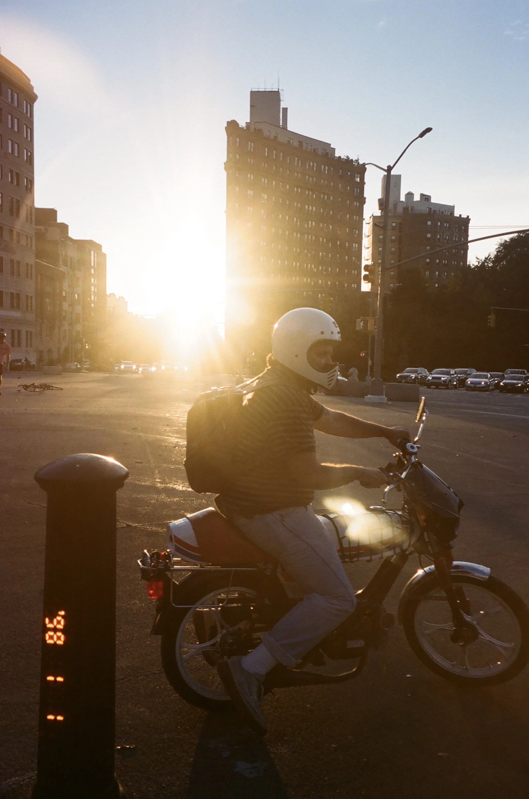 A person riding a motorcycle in an urban area during sunset, with tall buildings and a traffic light in the background.