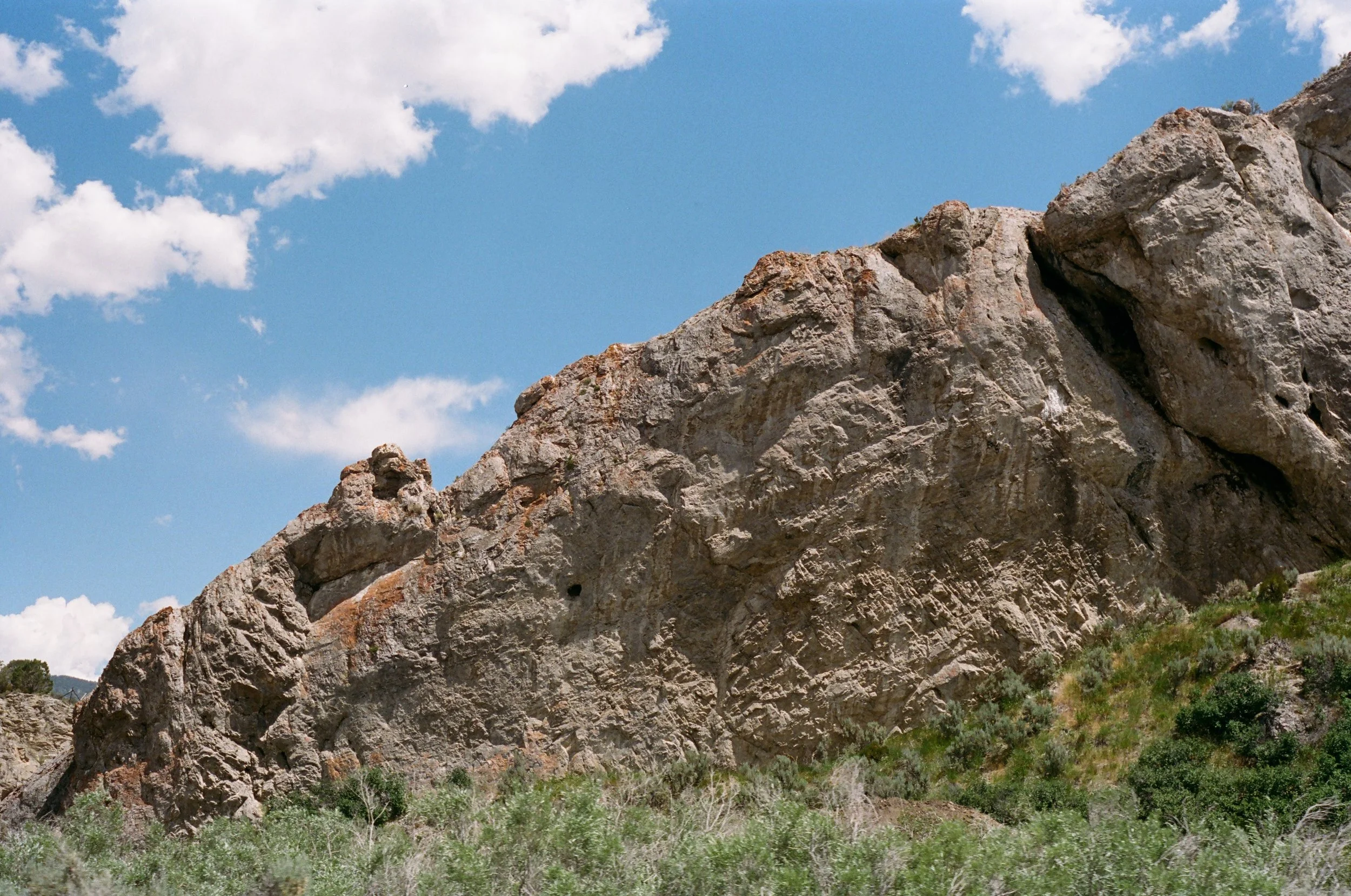 Large rocky mountain formation with steep, rugged cliffs under a blue sky with scattered white clouds, surrounded by green vegetation.
