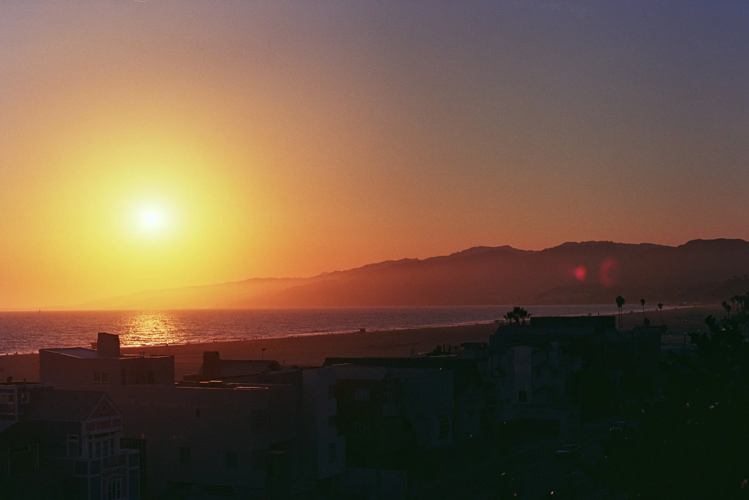 Sunset view over the ocean with silhouetted houses and palm trees in the foreground, and mountains in the distance.