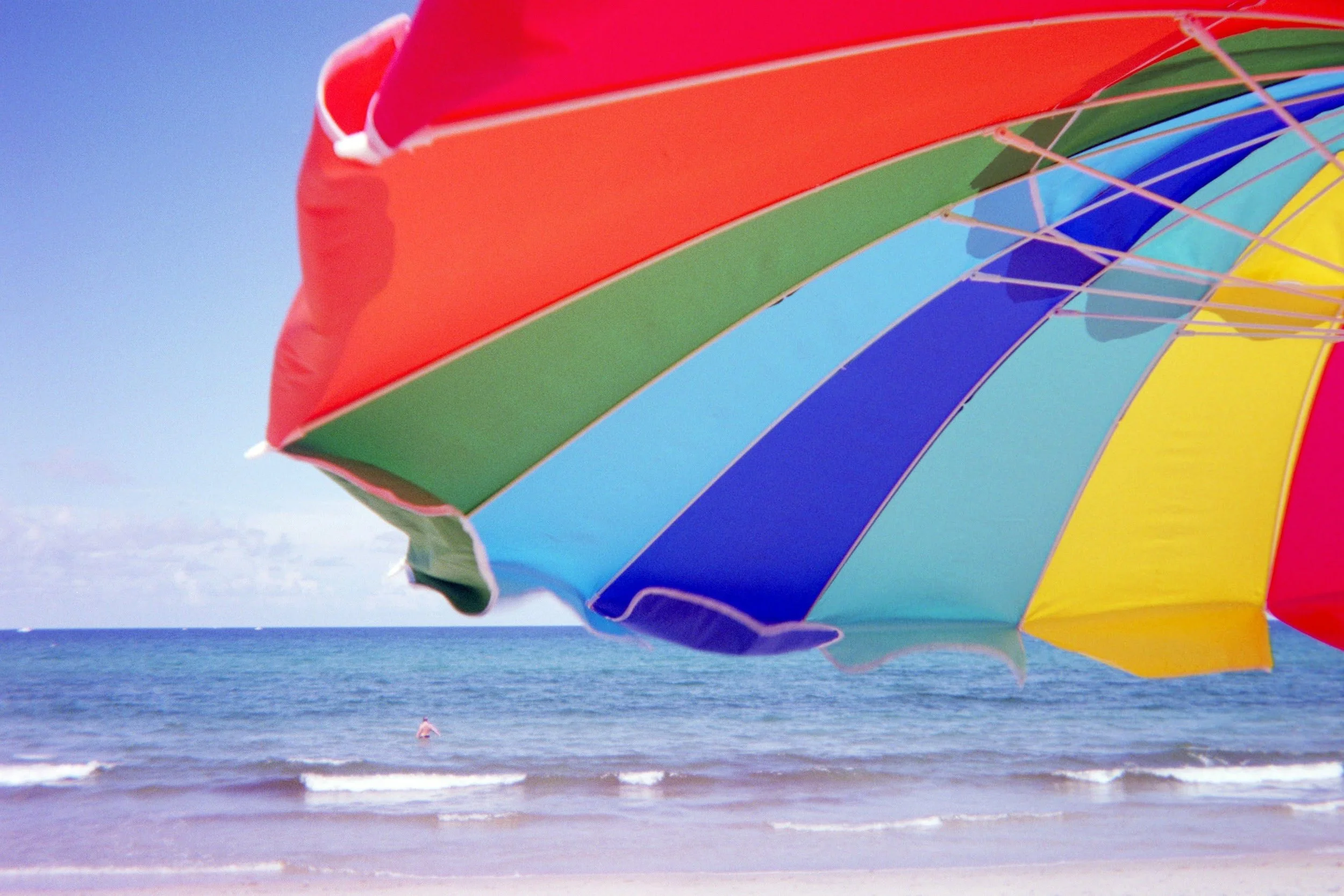 A large colorful beach umbrella with red, purple, green, blue, and yellow panels on the beach, with ocean waves and a person in the distance.