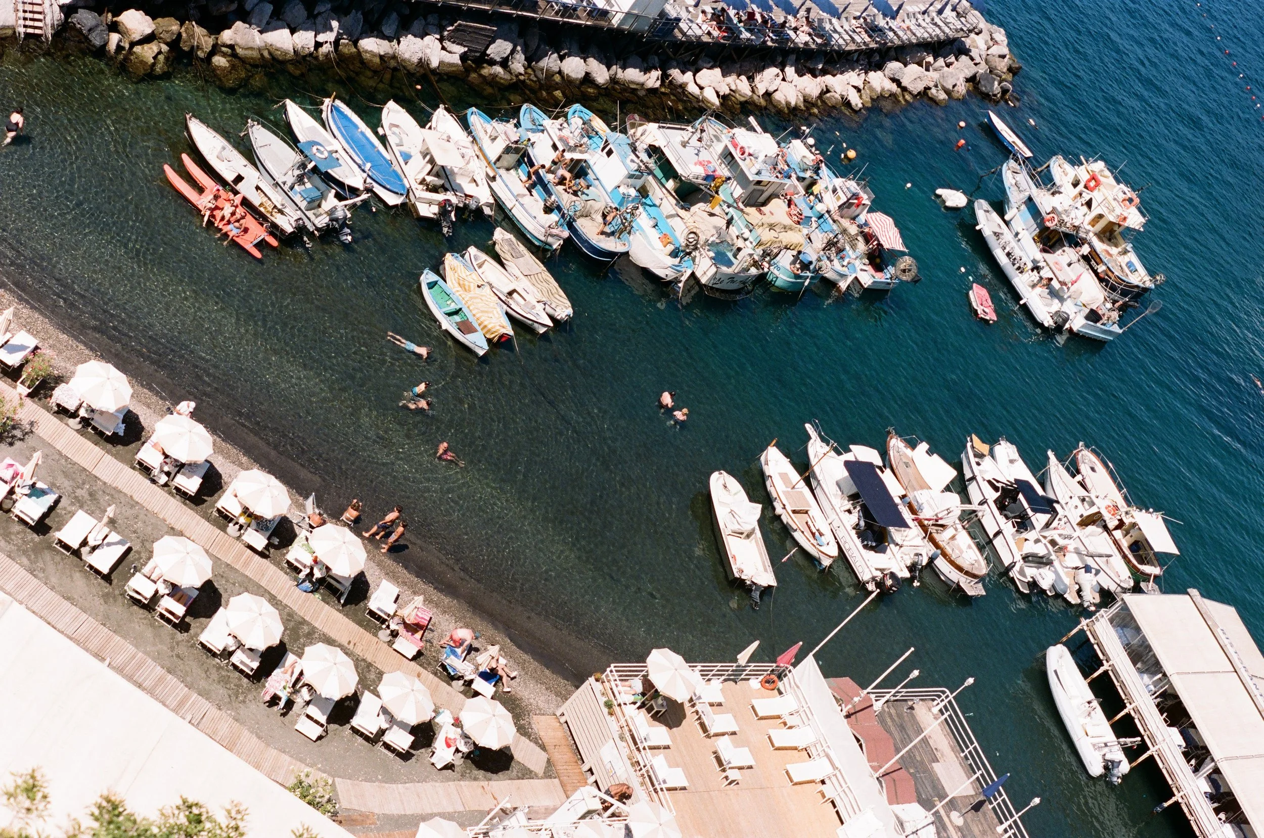 Aerial view of a harbor with boats docked along the pier, people swimming and sunbathing, and umbrellas and lounge chairs on the beach.