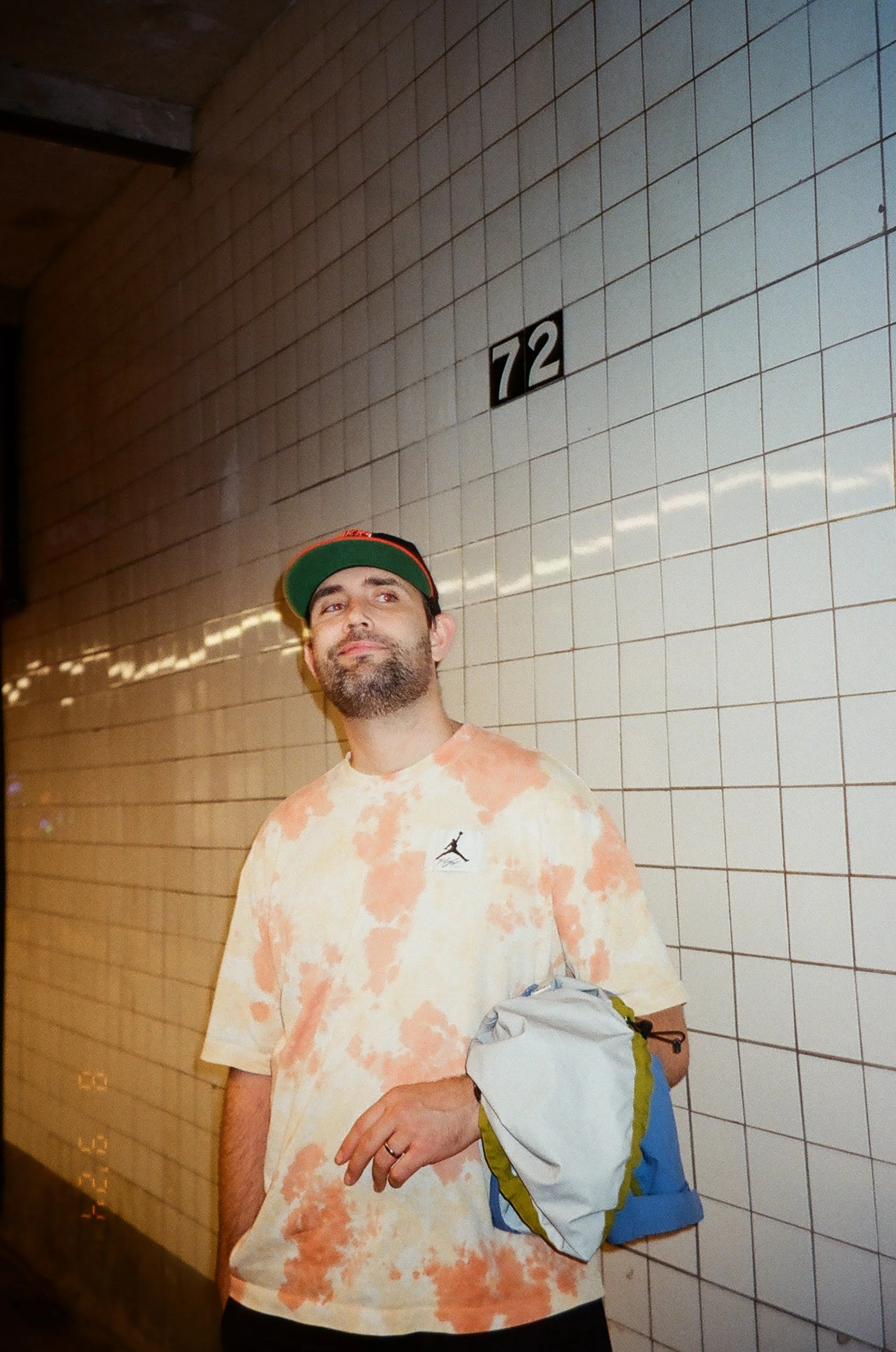 A man with a beard and mustache wearing a red and green baseball cap, a tie-dye t-shirt, and holding a white and blue jacket, standing against a tiled wall with the number 72 displayed above him.