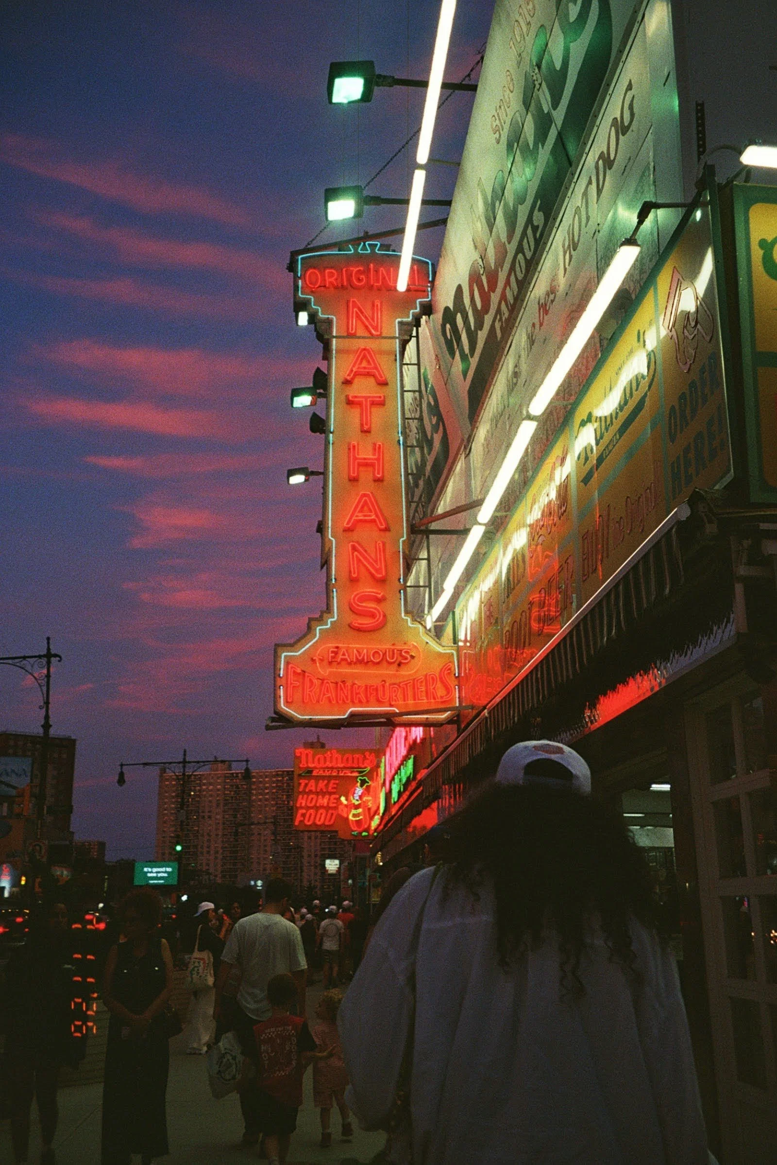 A lively city street scene during dusk with neon signs, including a large vertical sign for Nathan's Famous Hot Dogs and various other illuminated signs advertising food and takeout options, with a crowd of people walking and standing along the sidew