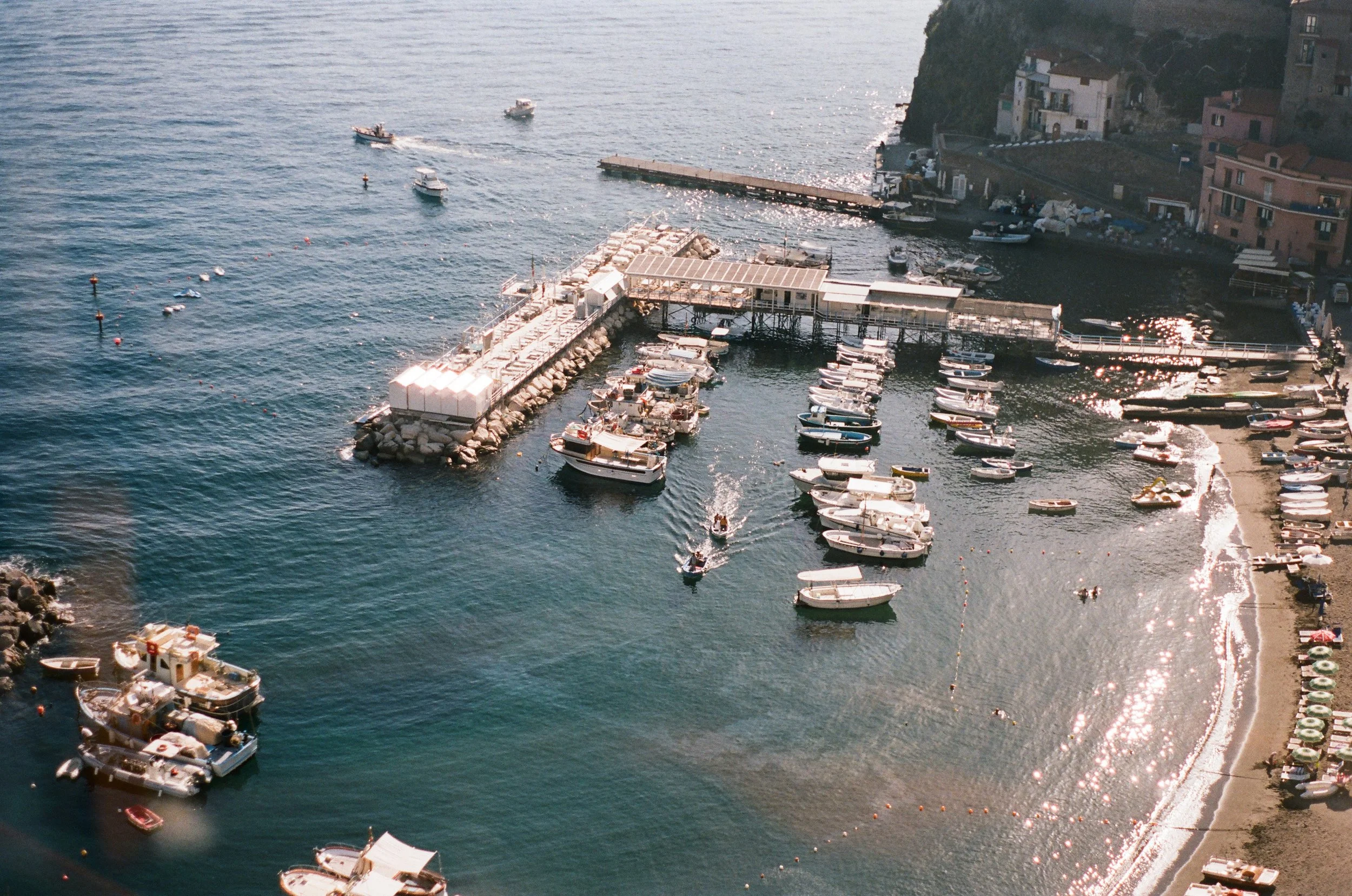 A seaside harbor with multiple boats docked and sailing, a pier extending into the water, and colorful buildings on a cliffside in the background.