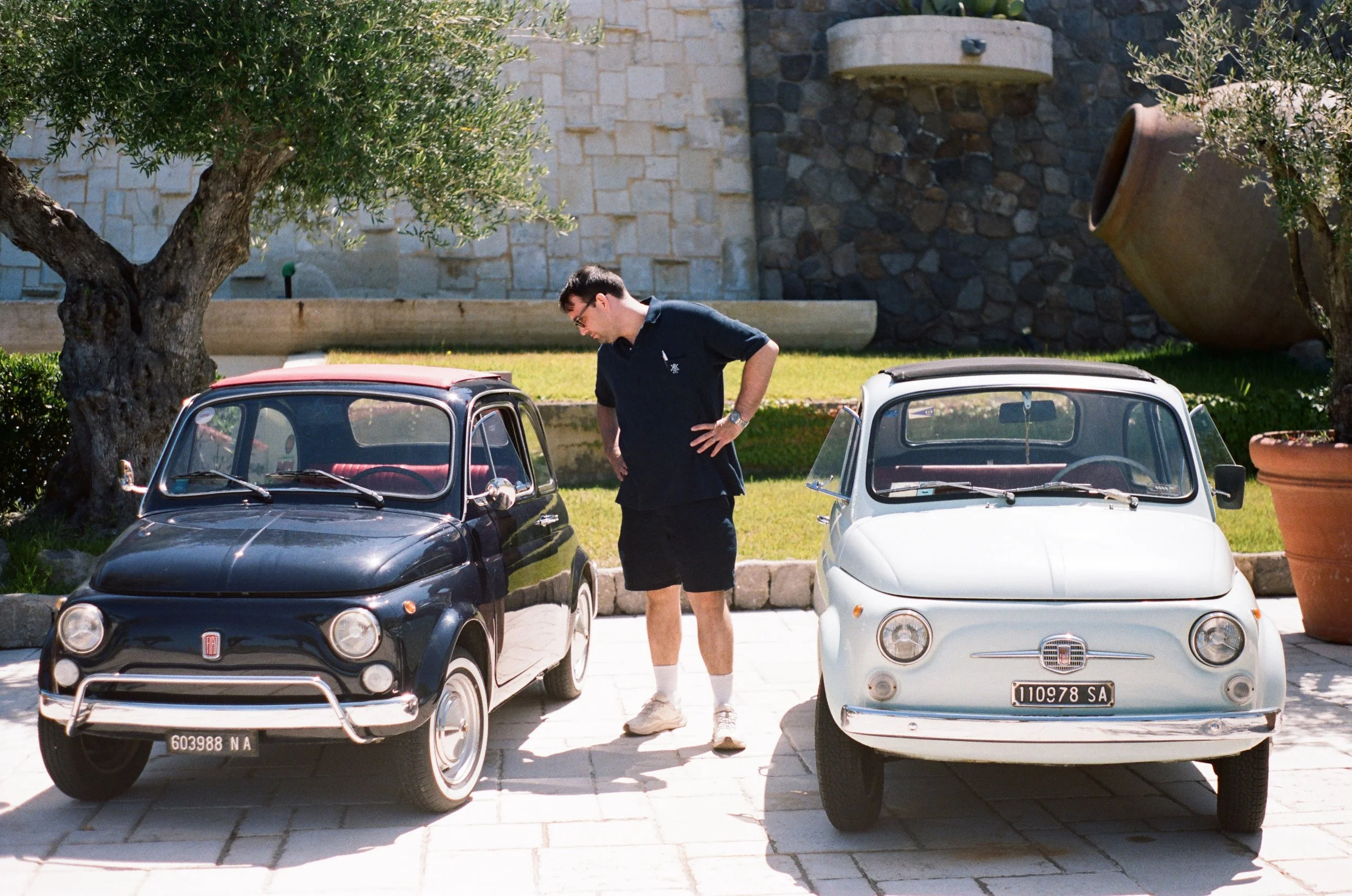 A man wearing glasses, a black polo shirt, shorts, and sneakers stands between two vintage Fiat 500 cars, looking into the black one.