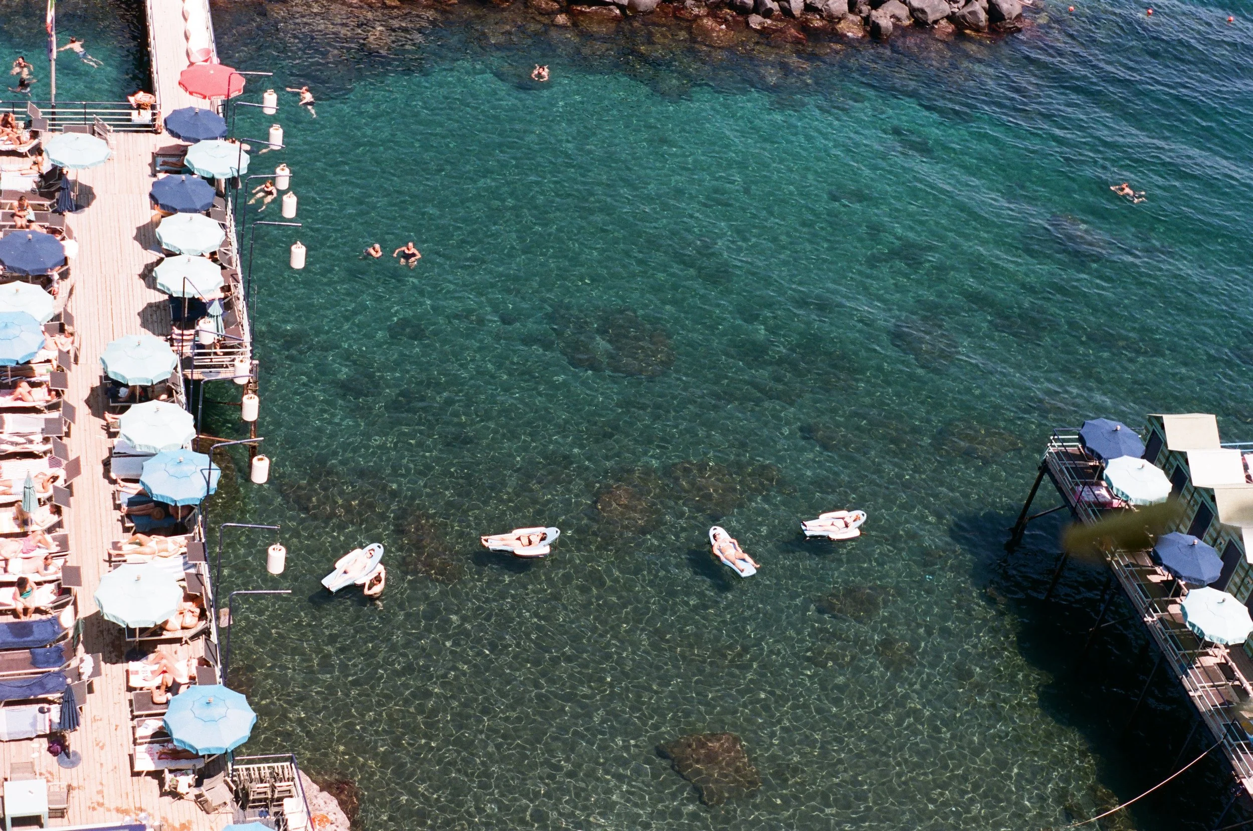 Aerial view of a crowded seaside pier with blue umbrellas, lounge chairs with sunbathers, swimmers in the clear water, small boats, and rocky shoreline.