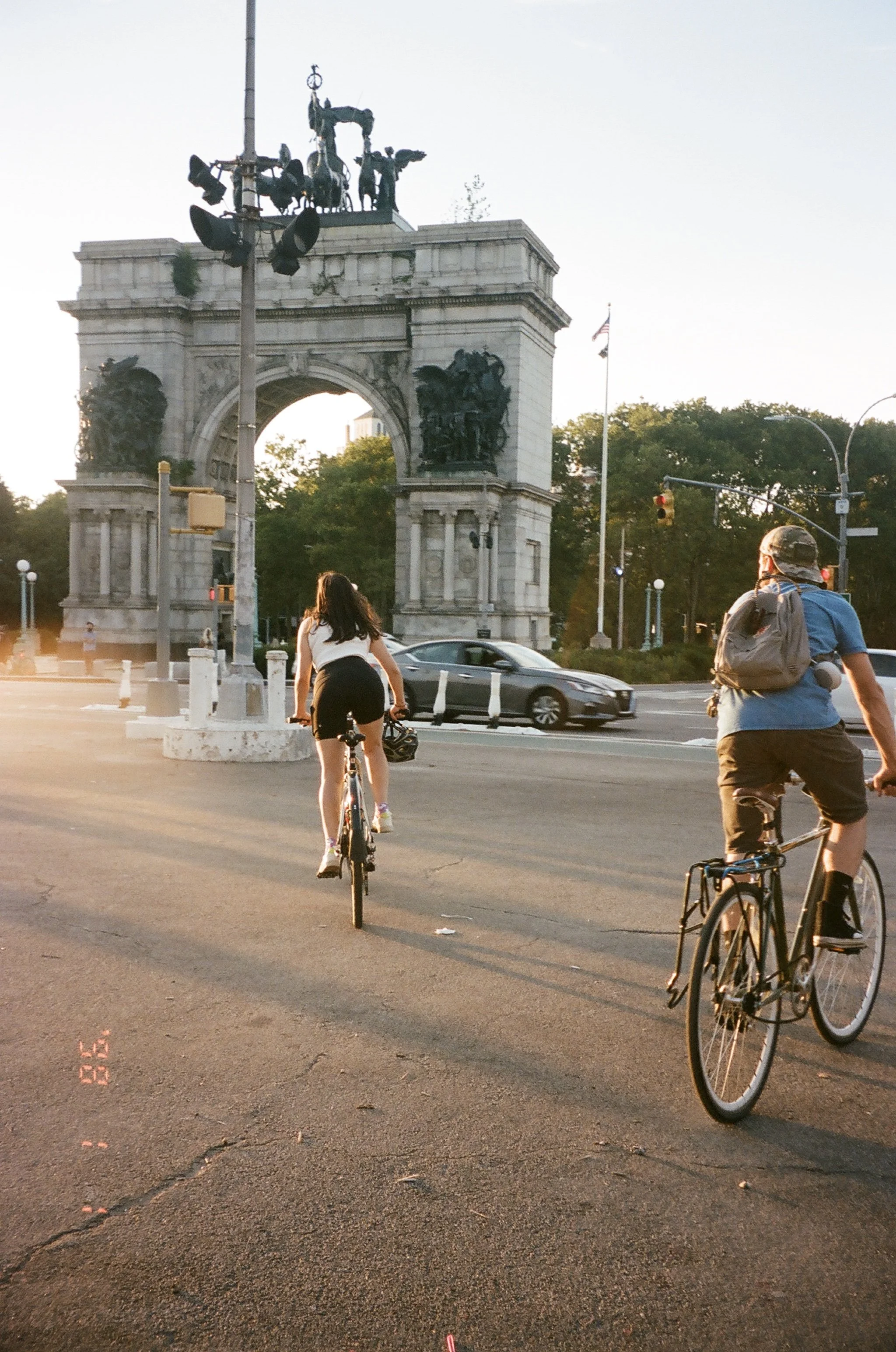 People riding bicycles near a historic arch monument in a city with greenery in the background during sunset.