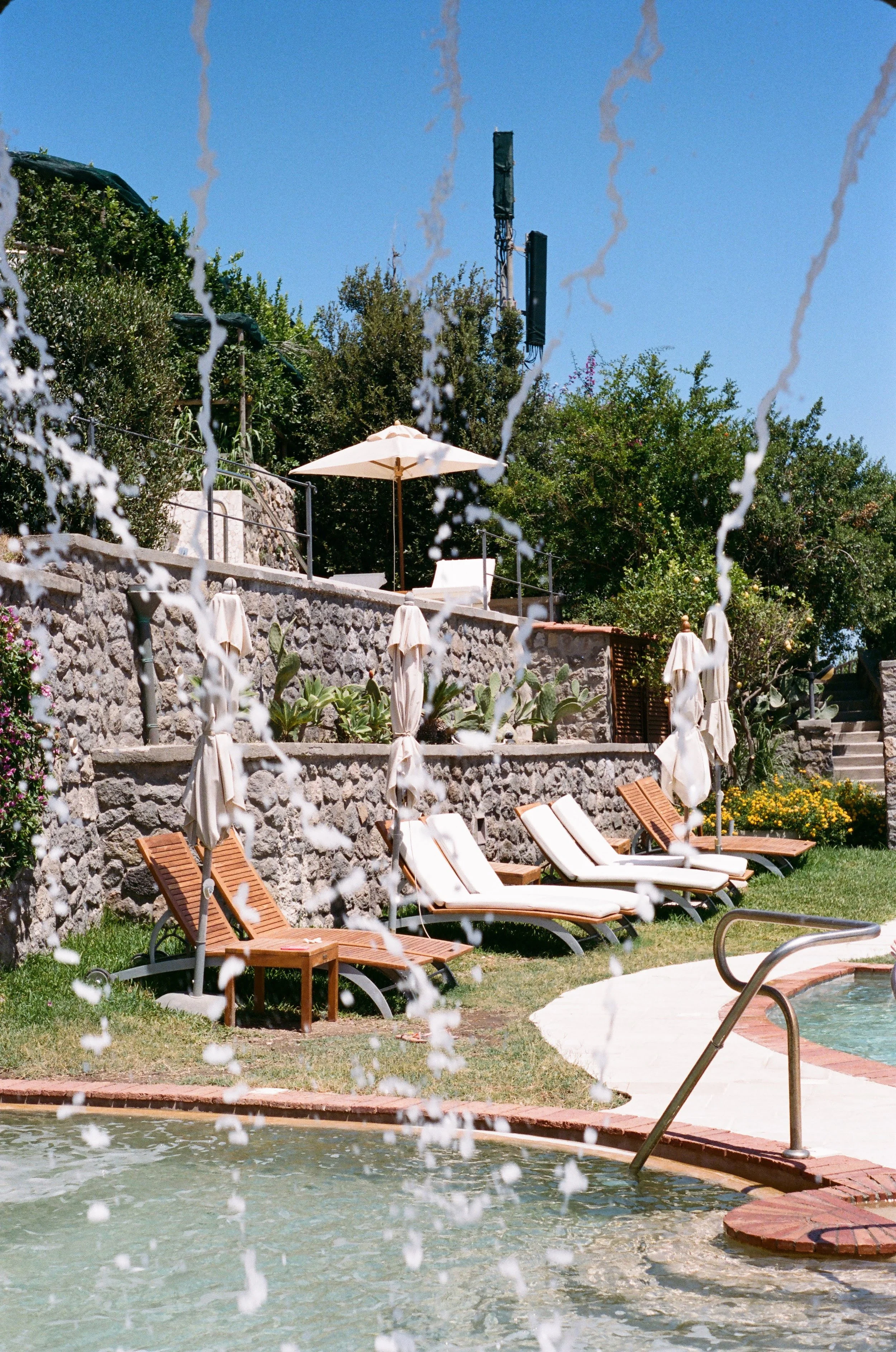Poolside scene with lounge chairs, umbrellas, and a water fountain, surrounded by green trees and a stone wall, under a clear blue sky.