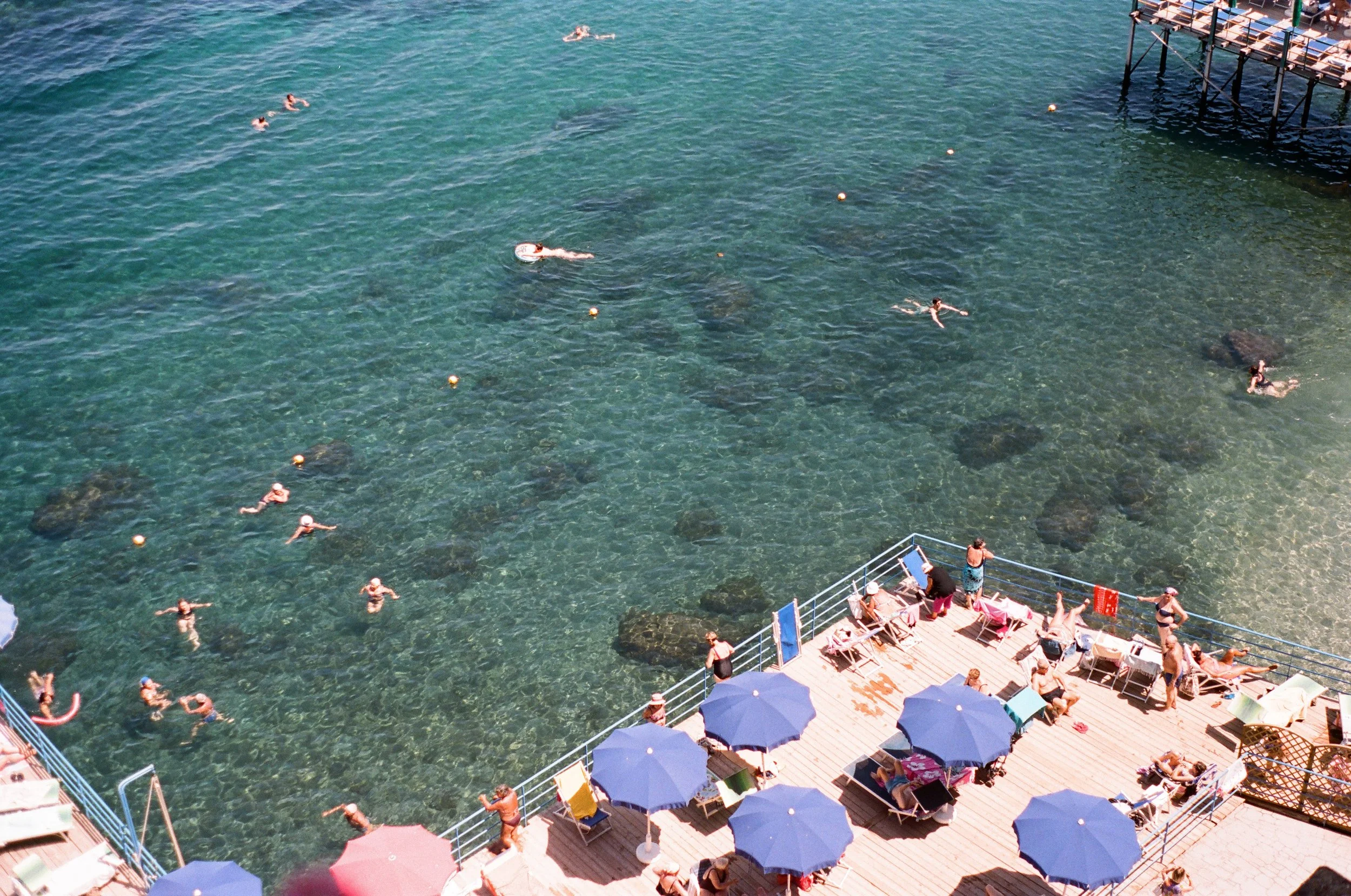 People swimming and lounging on a wooden pier at a beach with umbrellas and sunbathers, and a rocky shoreline beneath clear turquoise water.