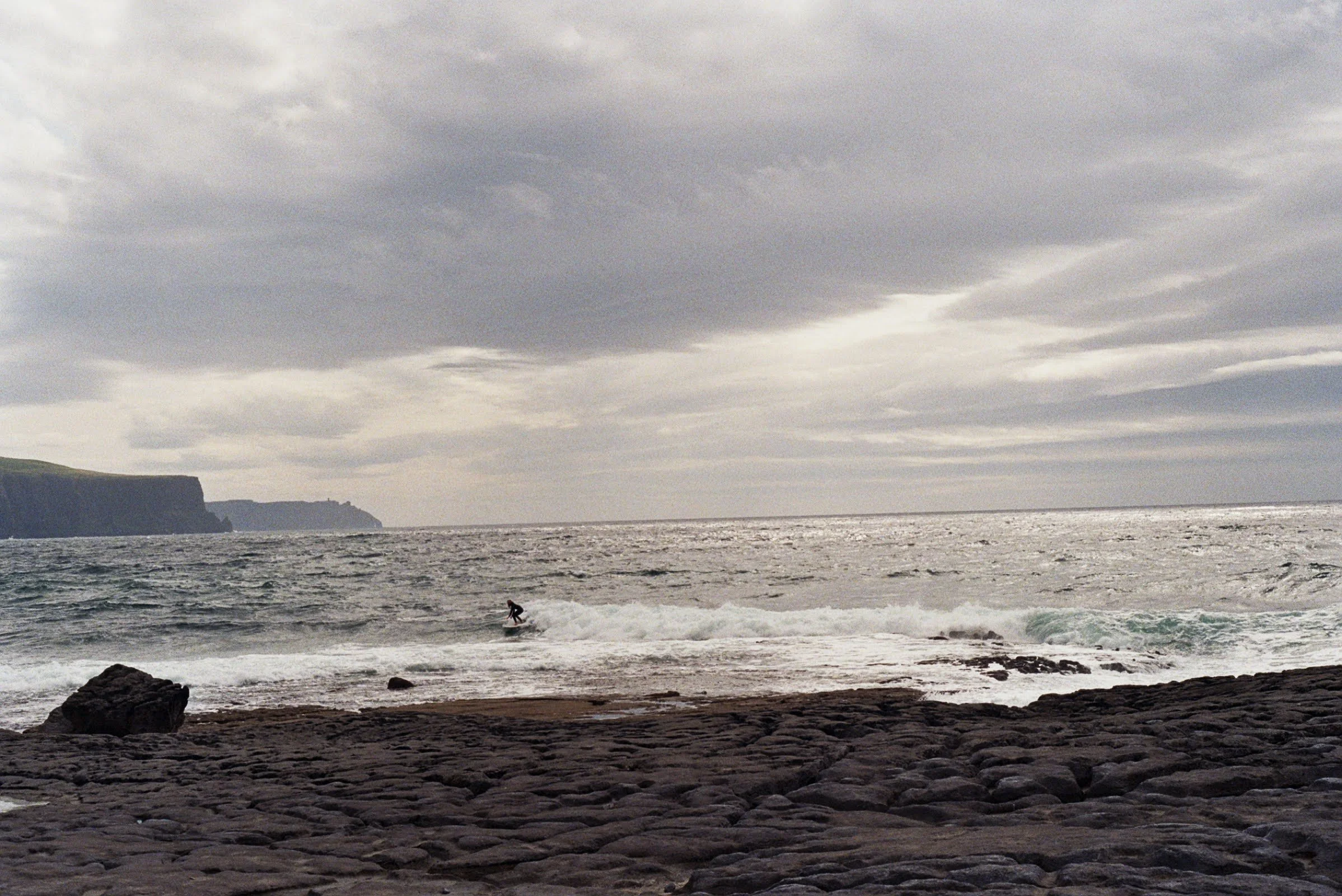 Overcast sky over ocean with a surfer riding a wave near rocky shoreline.