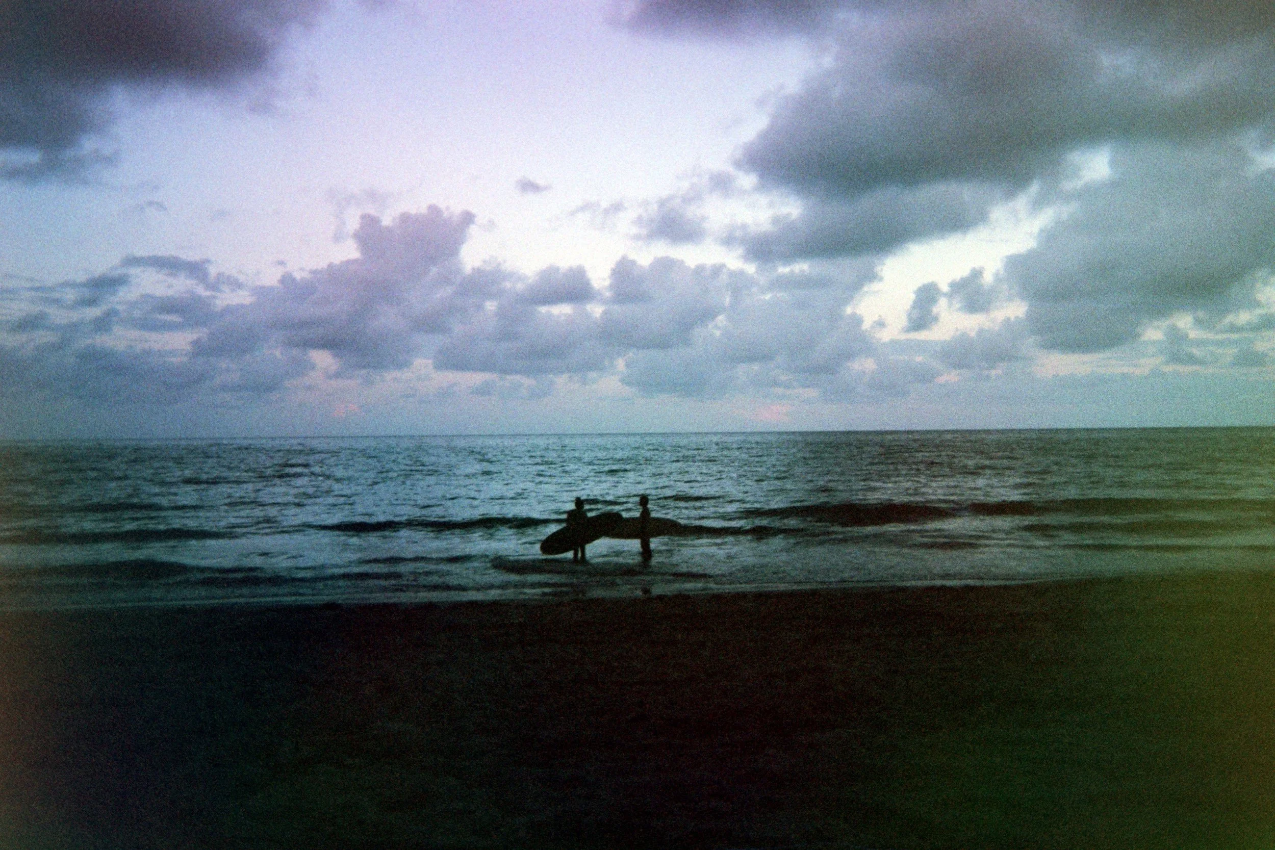 Two people with surfboards standing on the beach near the water, looking out at the ocean under a cloudy sky.