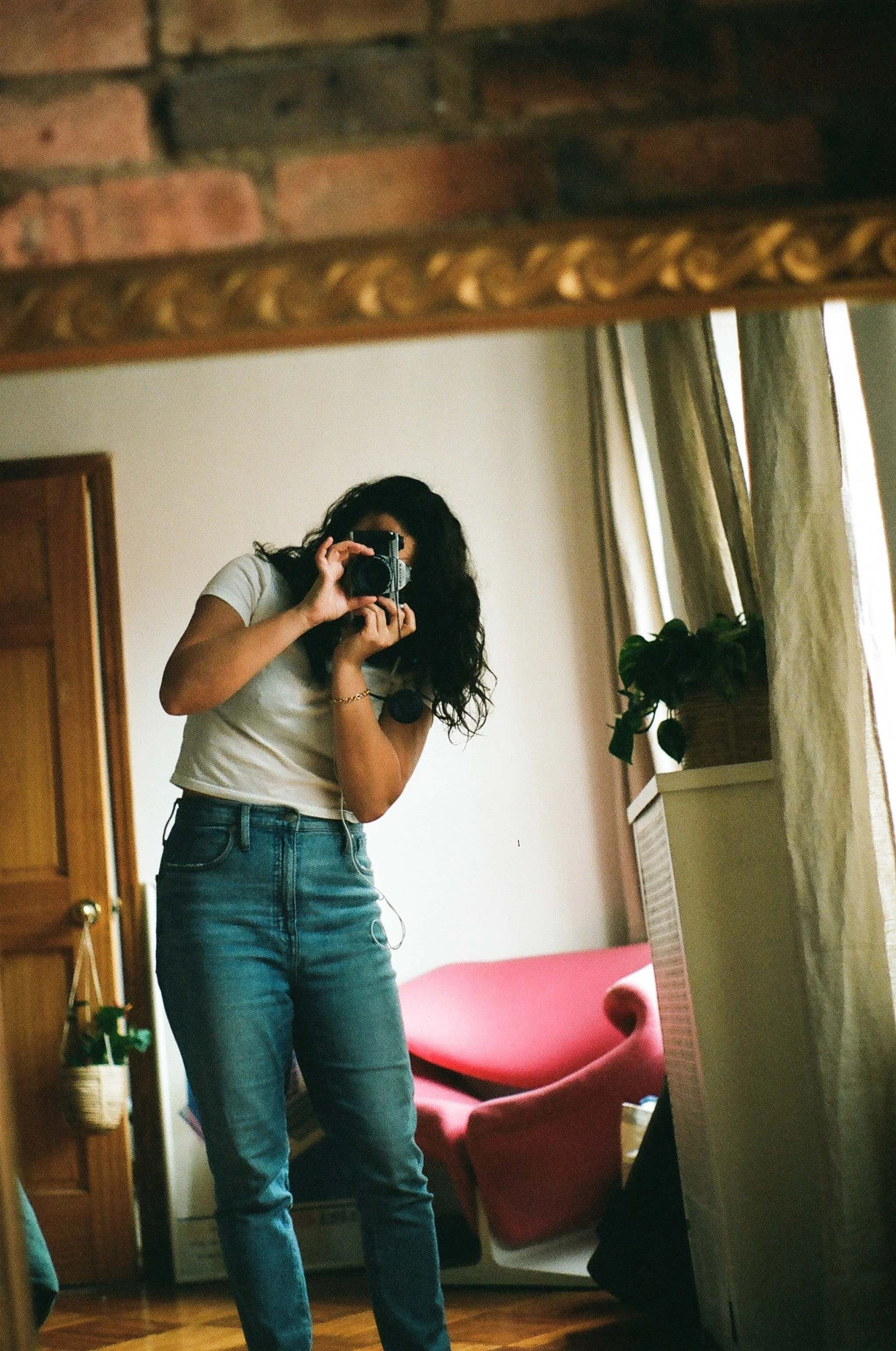 A woman with curly hair taking a mirror selfie with a camera in a room featuring wooden furniture, a red sofa, and potted plants.