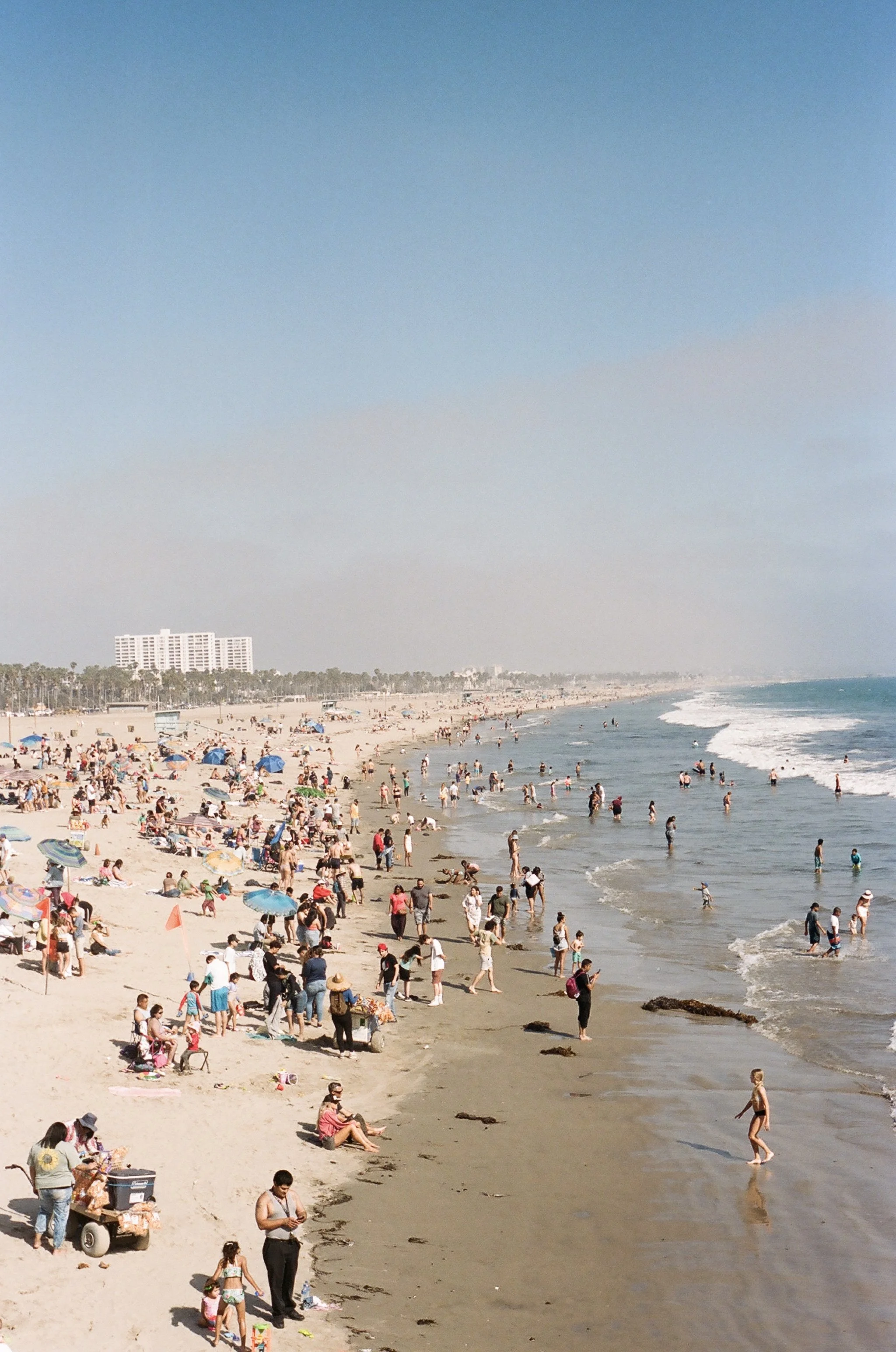 Crowded beach scene with many people swimming, sunbathing, and walking along the shoreline. Colorful umbrellas scattered across the sand and a large white hotel building in the background under a clear blue sky.