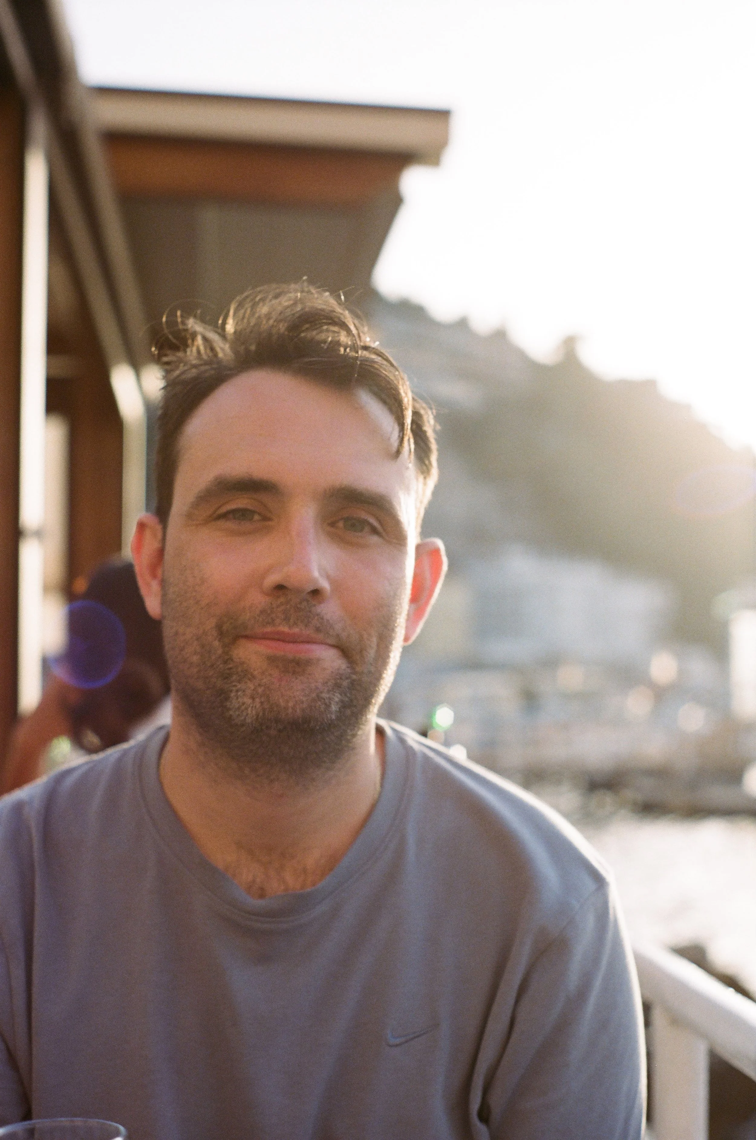 Close-up of a man with short dark hair and stubble, wearing a gray T-shirt, smiling outdoors during the daytime with sunlight in the background.
