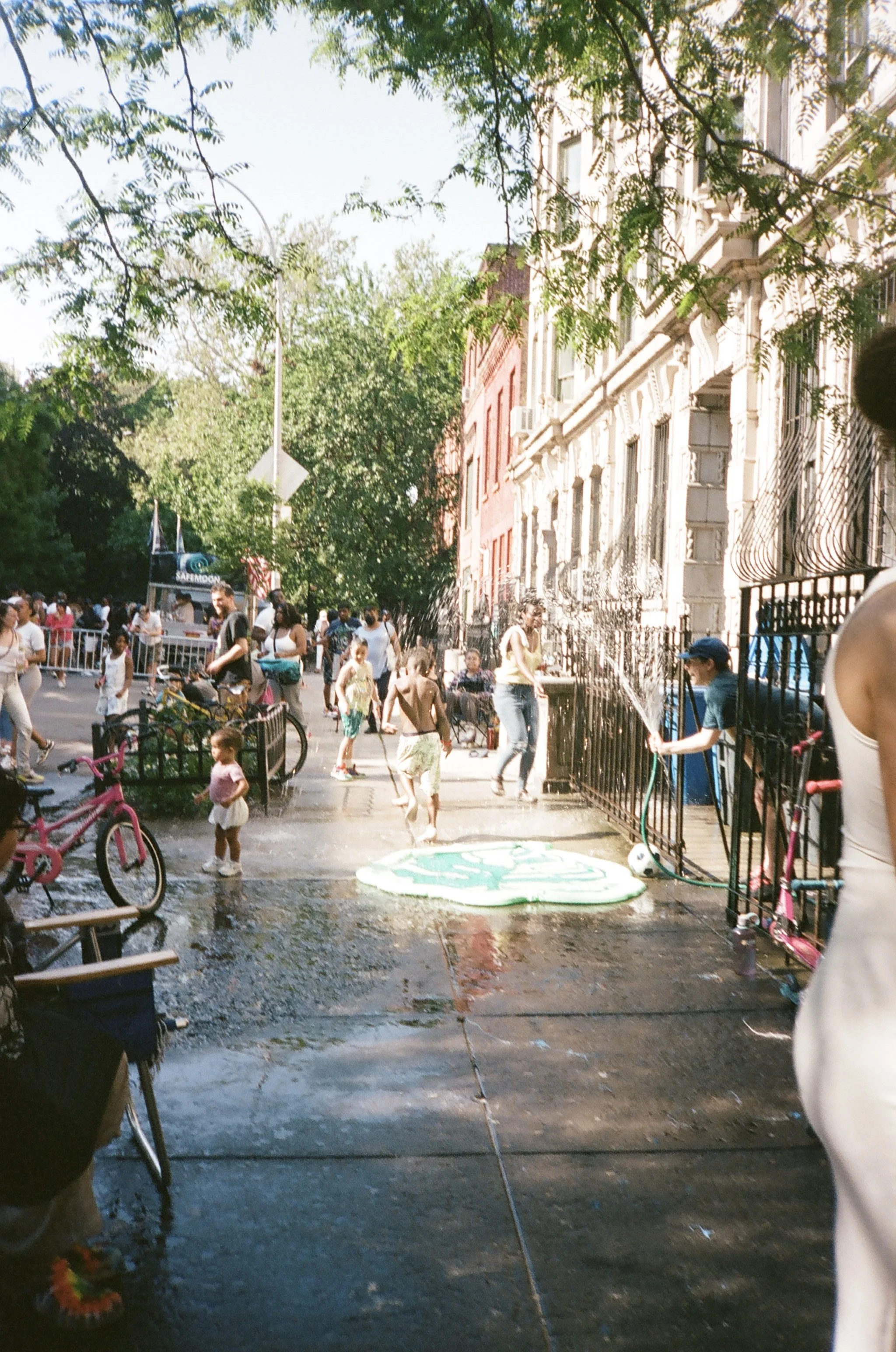 Street scene with people gathering, children playing, bicycles, and a water spray in a city neighborhood with brownstone buildings and trees.