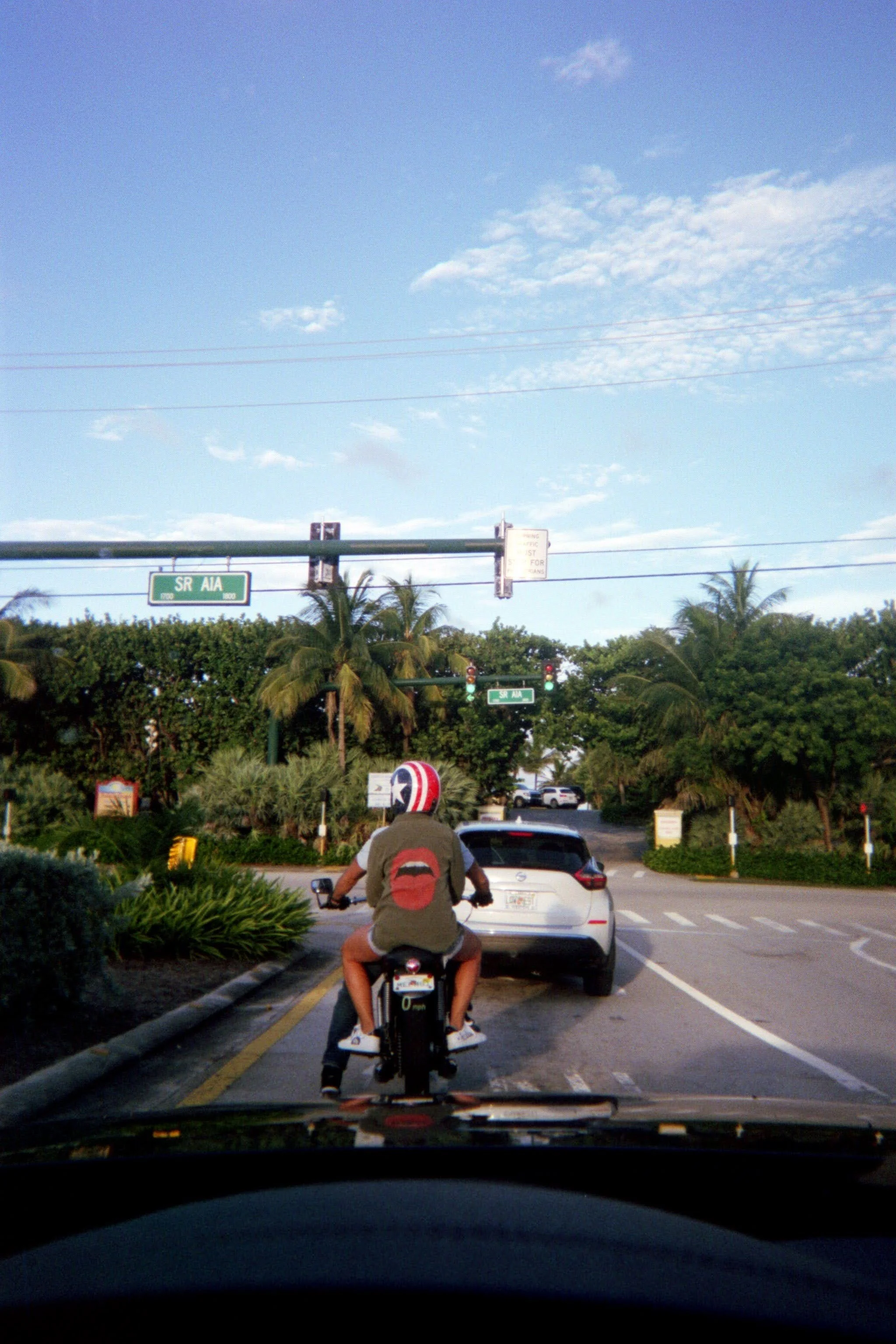 Motorcyclist riding in front of a white car at an intersection with traffic lights, surrounded by greenery and palm trees, under a blue sky with some clouds.