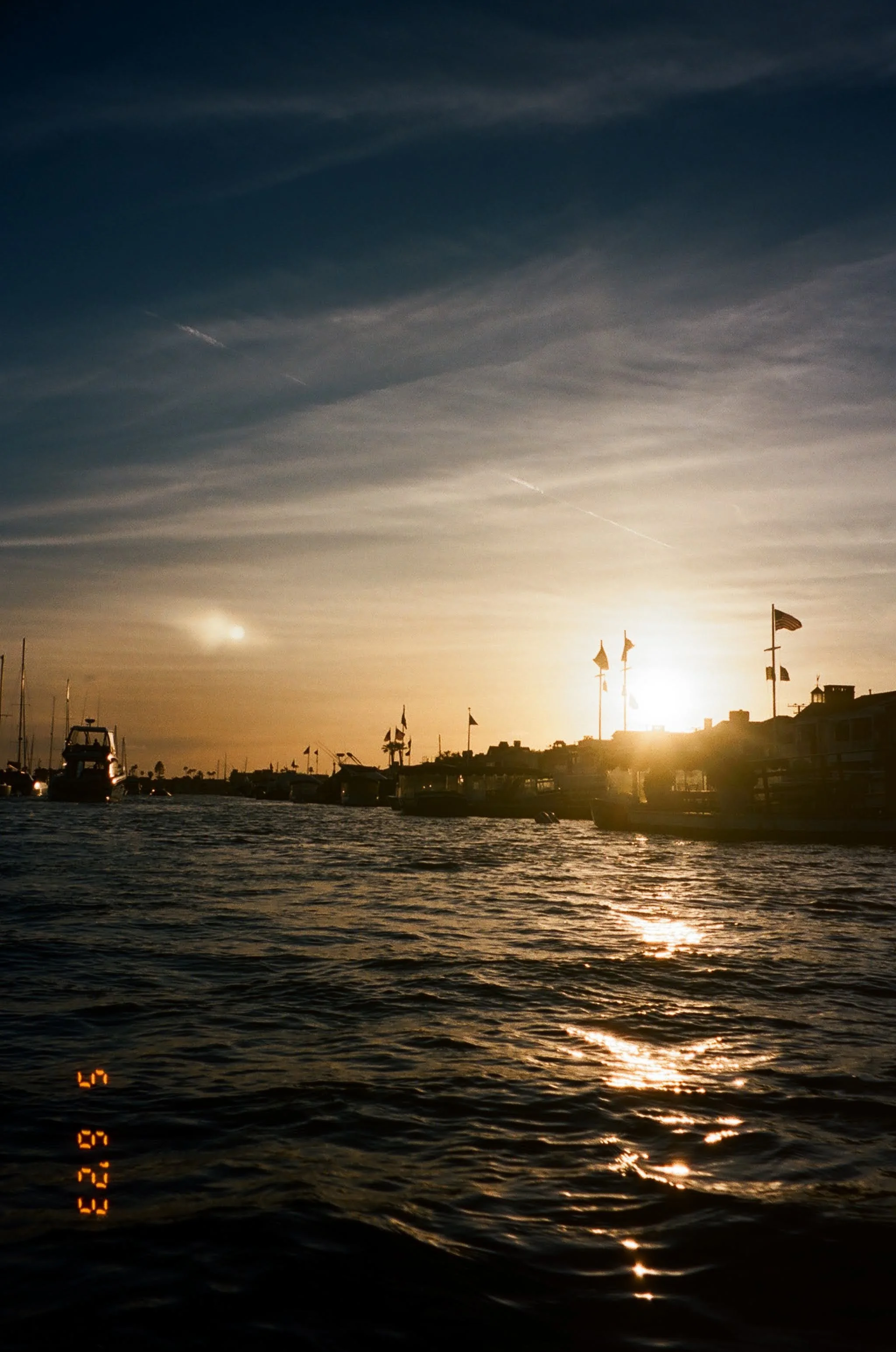 Sun setting over a harbor with boats and flags, creating a warm glow on the water surface.