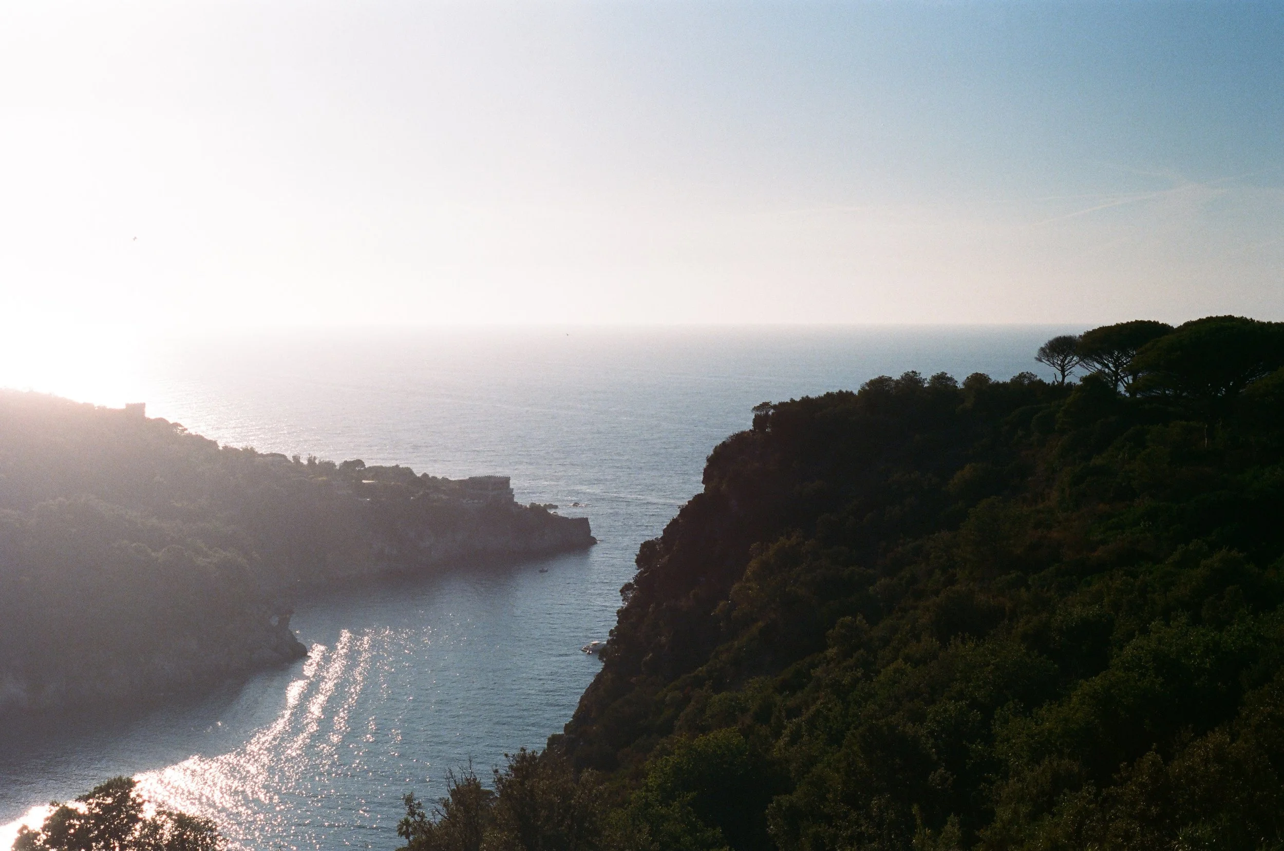 Scenic view of a coastal landscape with a fjord, surrounded by lush green hills and trees, with sunlight reflecting off the water during sunset or sunrise.