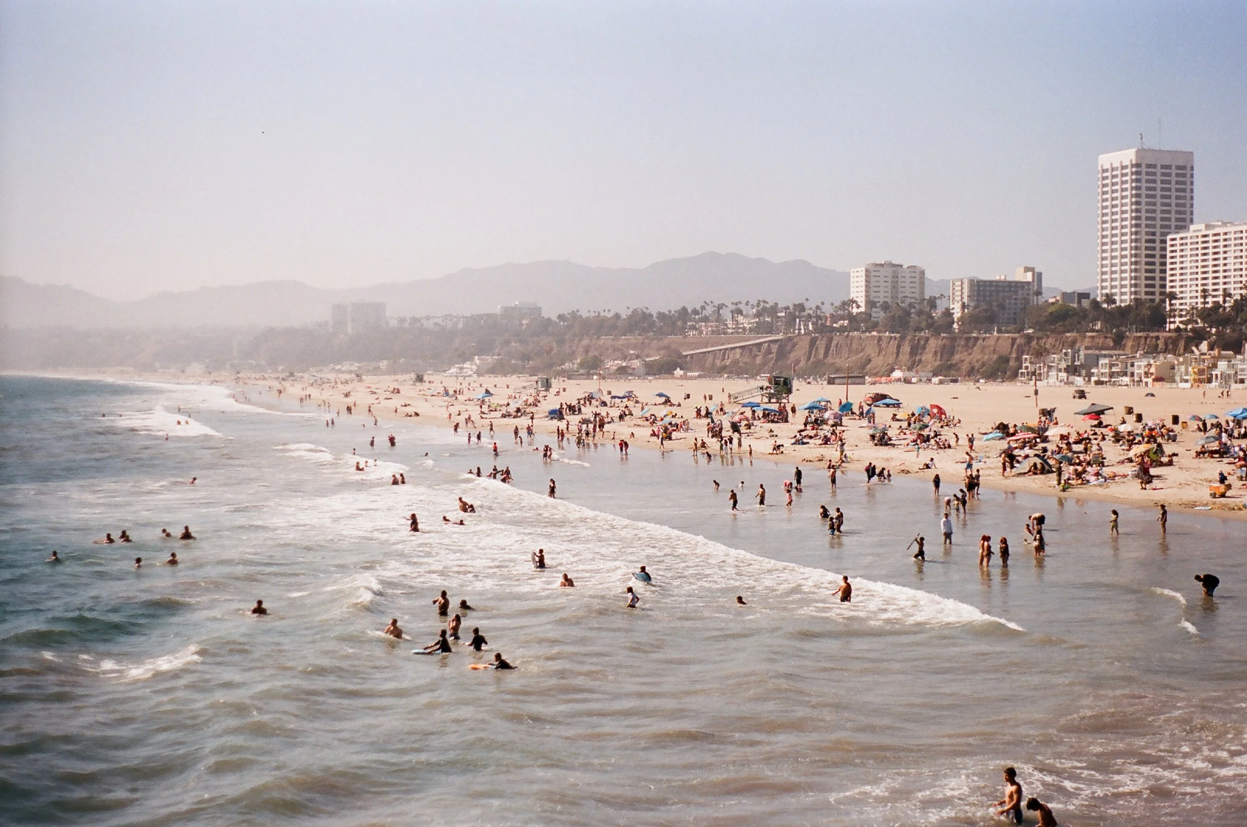 Beach scene with many people swimming and lounging under umbrellas, with high-rise buildings and mountains in the background.
