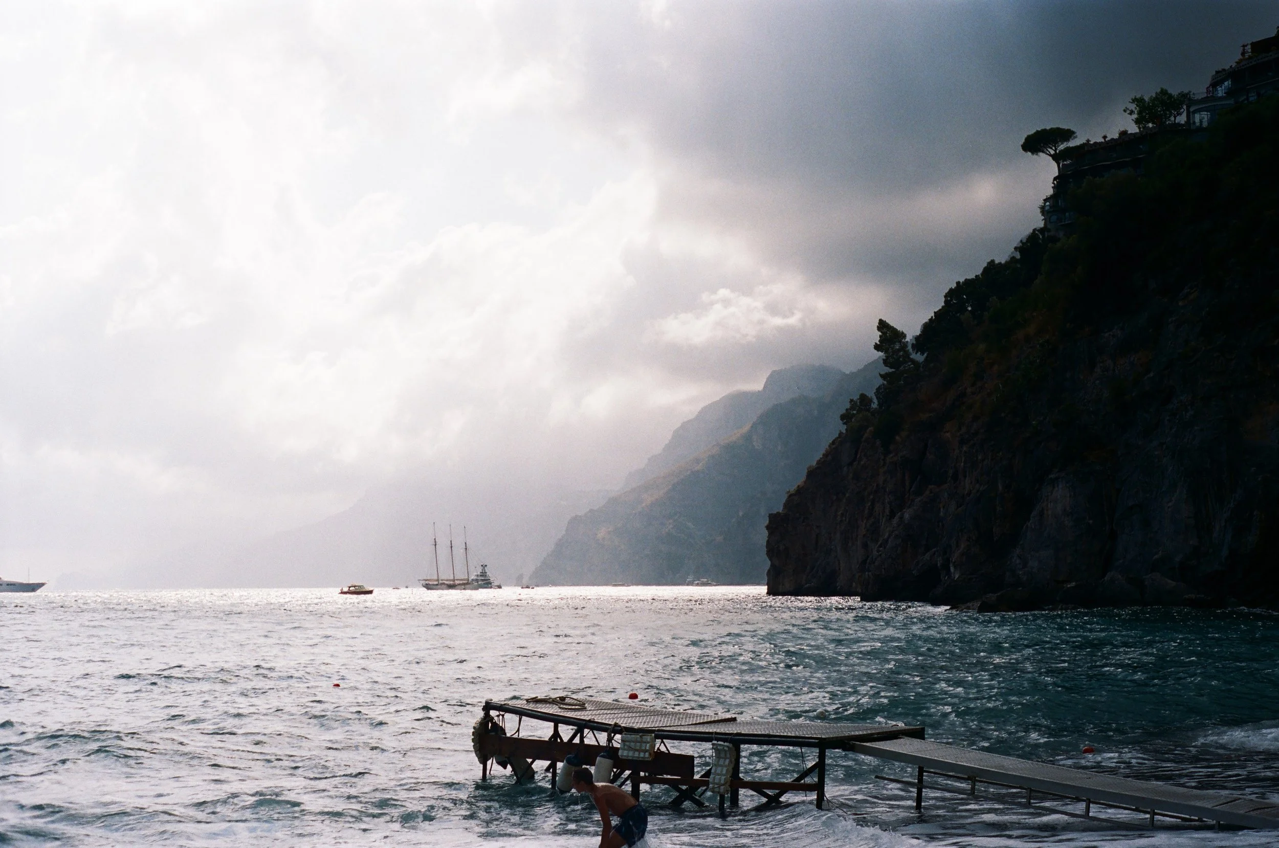 A cloudy day at a coastal location with boats on the water and a rocky hillside with buildings and trees.