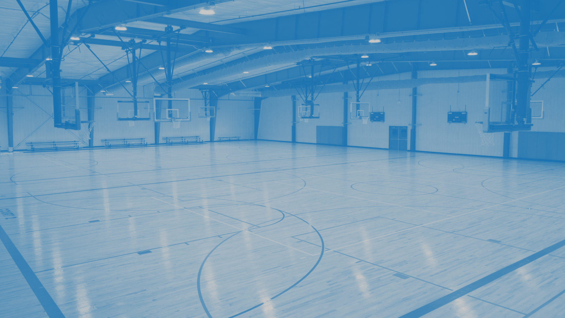 An indoor basketball gymnasium with hardwood floors, multiple basketball hoops, and high ceiling with lighting.