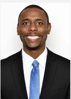 Headshot of a smiling man in a black suit, white shirt, and blue tie against a plain background.