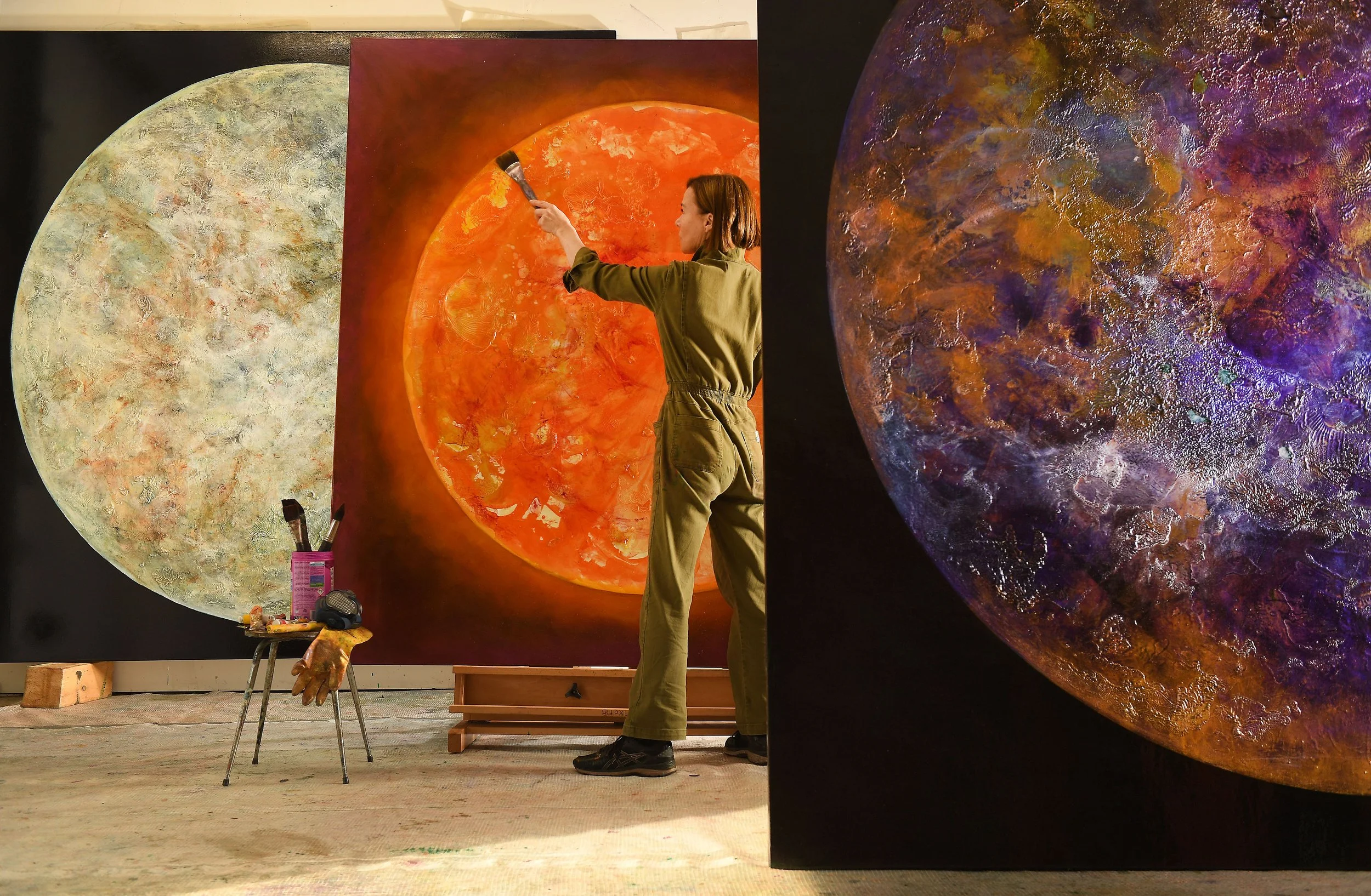 An artist painting a large, orange, sun-like artwork on a canvas. There are other large abstract art pieces visible in the background, with the artist standing in an art studio.