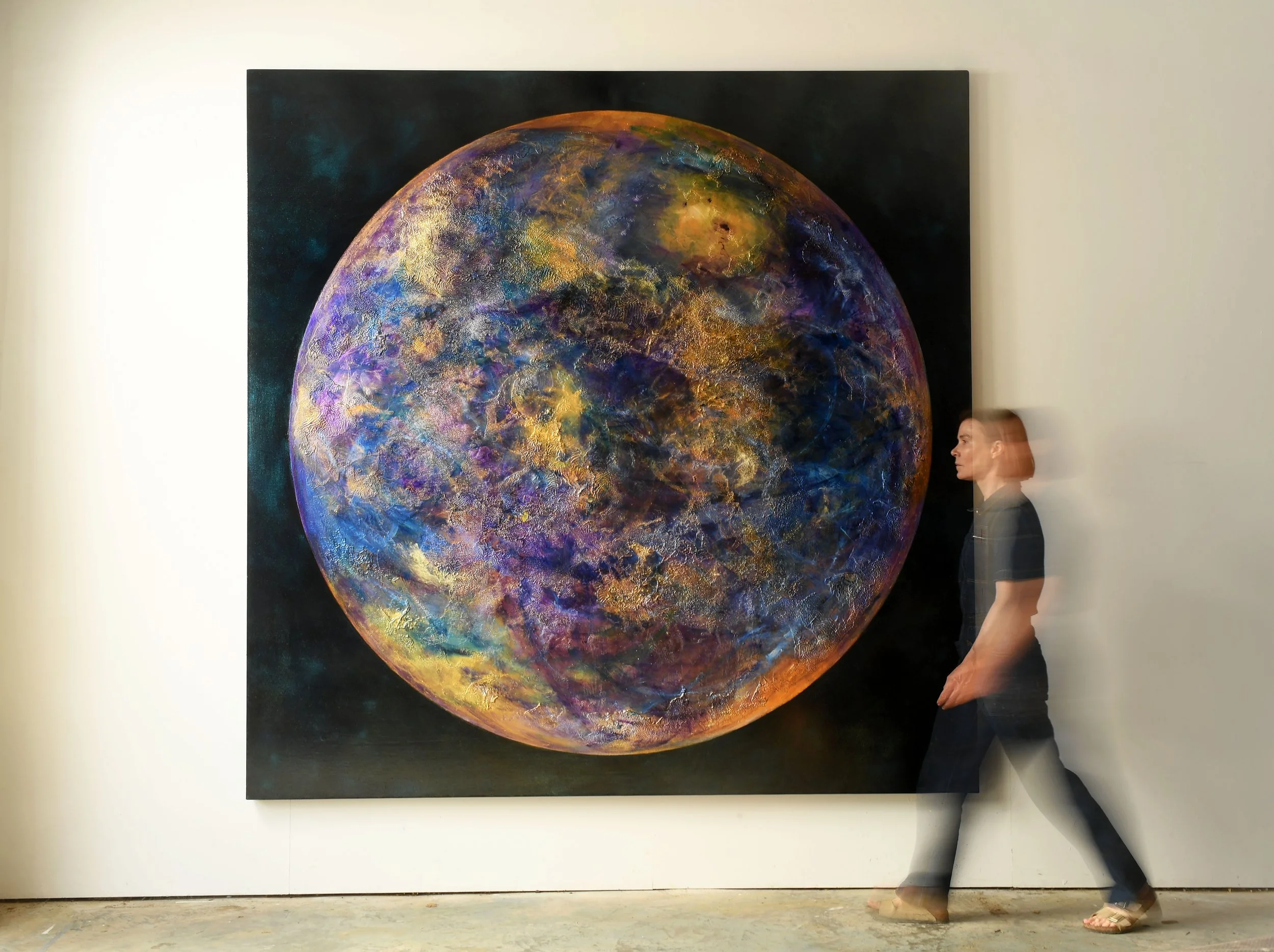 Large abstract painting of a colorful, textured planet or moon on a dark background, with a person standing and walking past it.