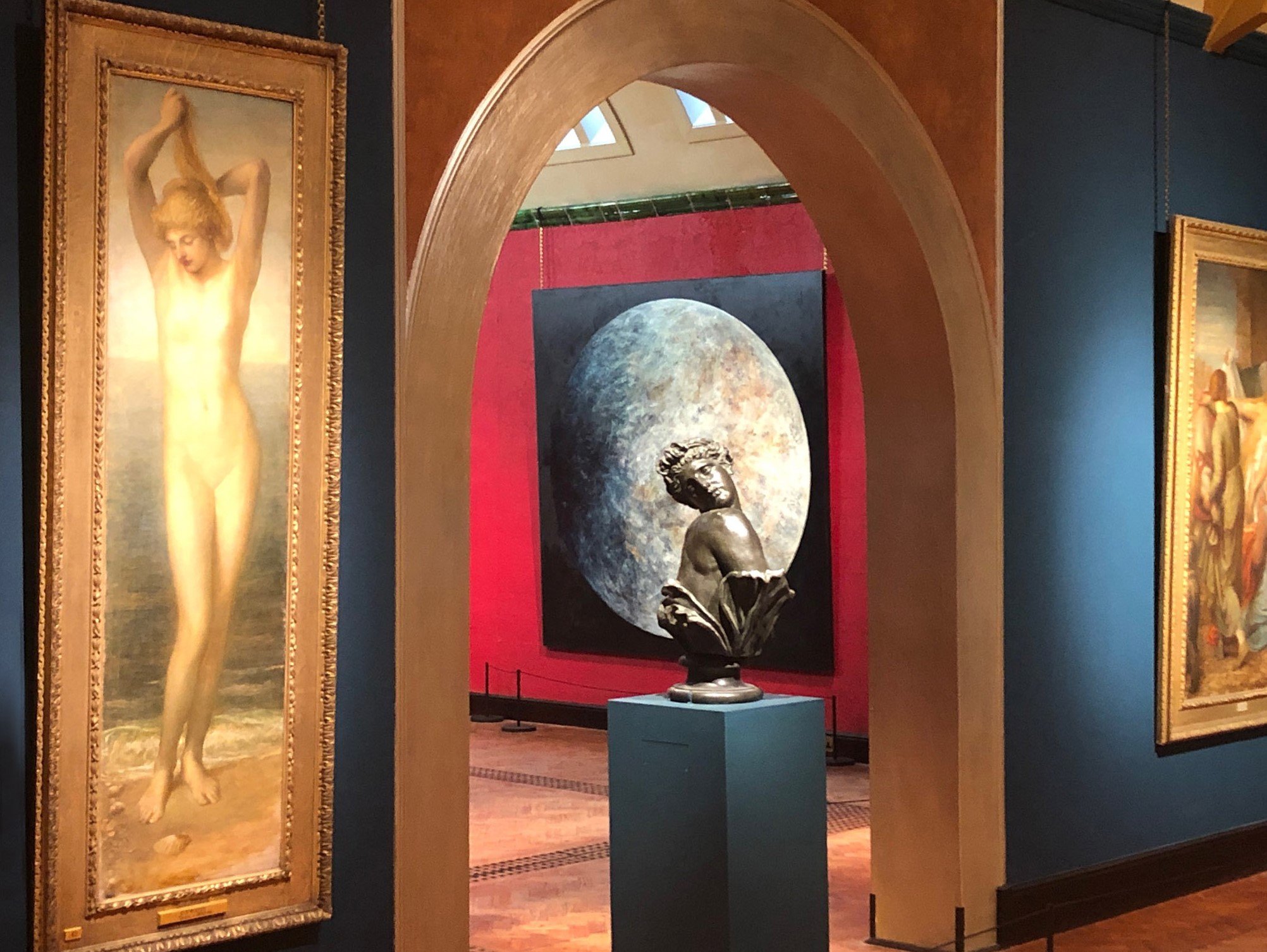 Art gallery featuring classical sculptures and paintings, including a large painting of a nude woman on the left and a sculpture of a seated woman with a moon in the background in the center.
