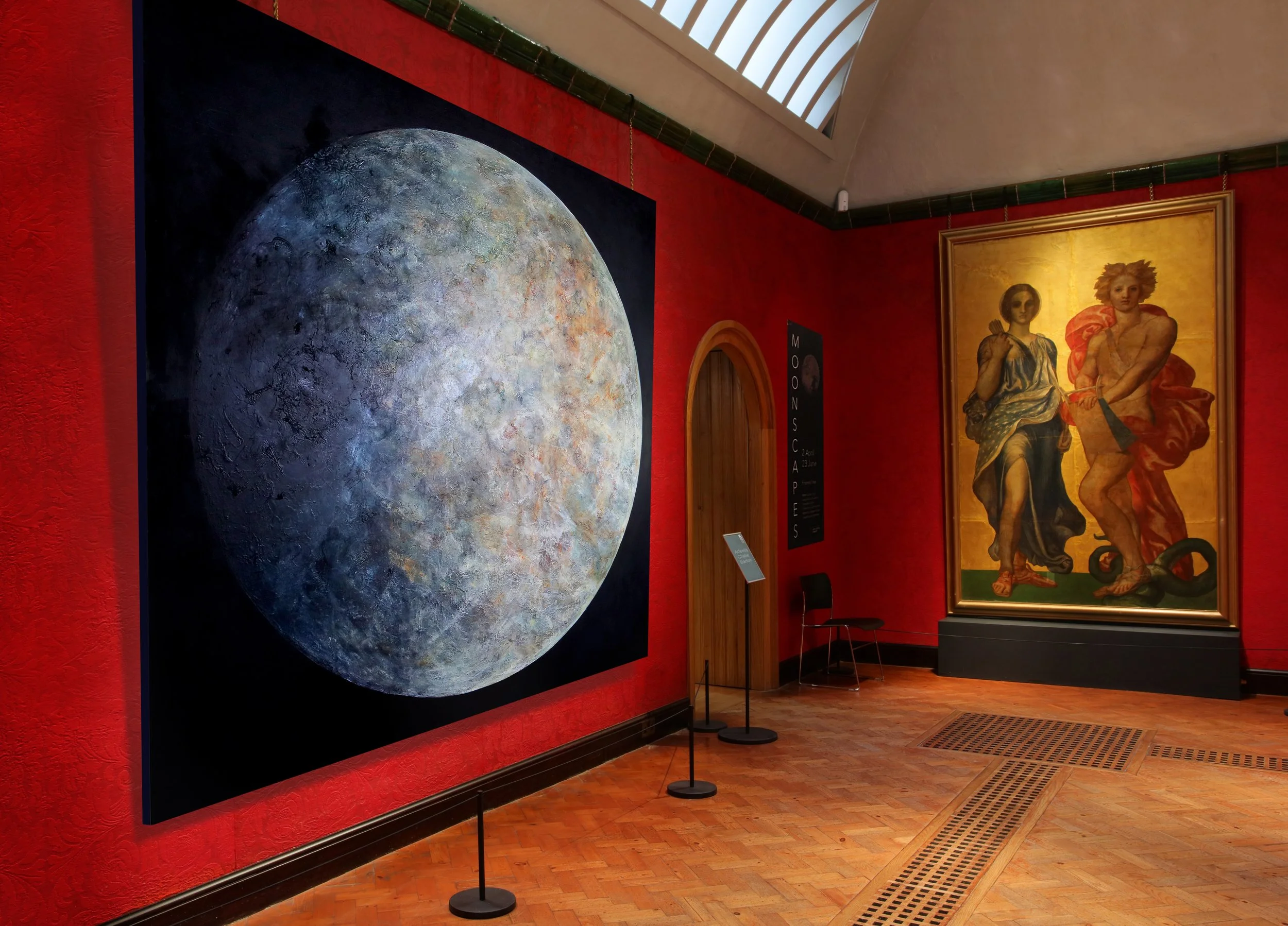 A gallery wall displaying a large photograph of the moon's surface on a black background, next to a framed painting of two women in classical attire on a golden background, in a room with red walls, a skylight, wooden floors, and small black signs with white text.